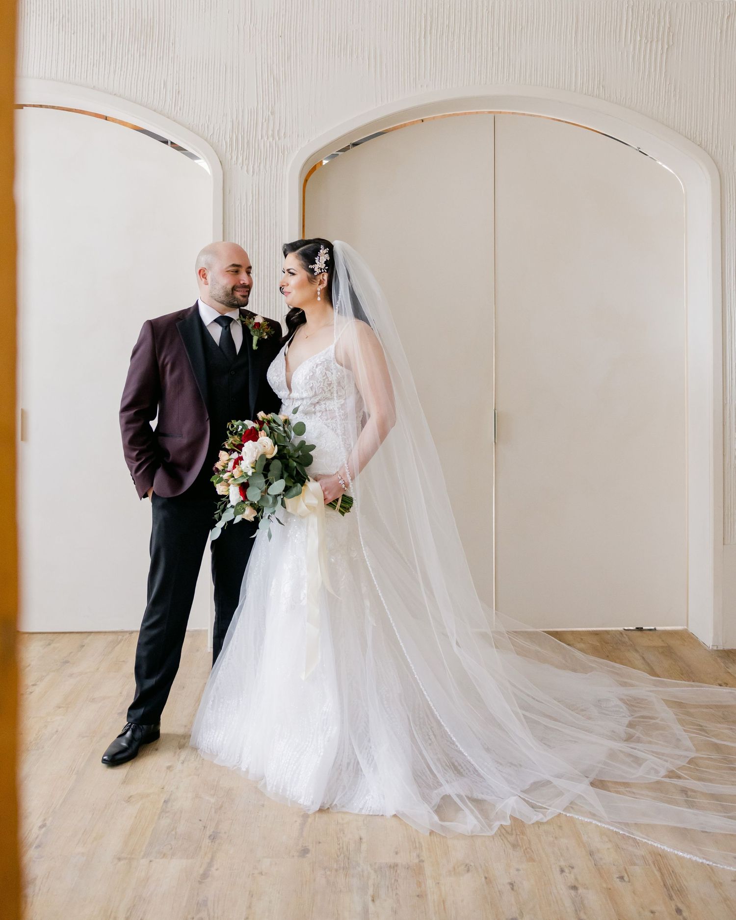 Wedding couple stands in elegant white room with arched doorways and wooden floors.