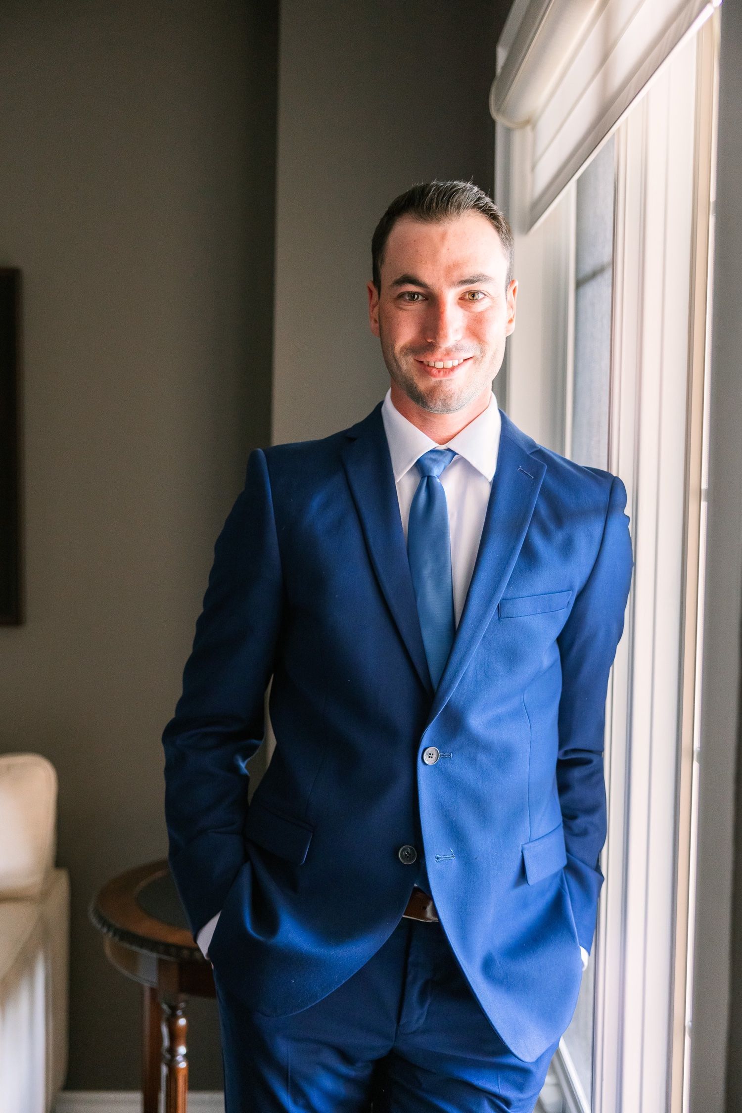 A dapper gentleman in a royal blue suit poses confidently by a bright window.