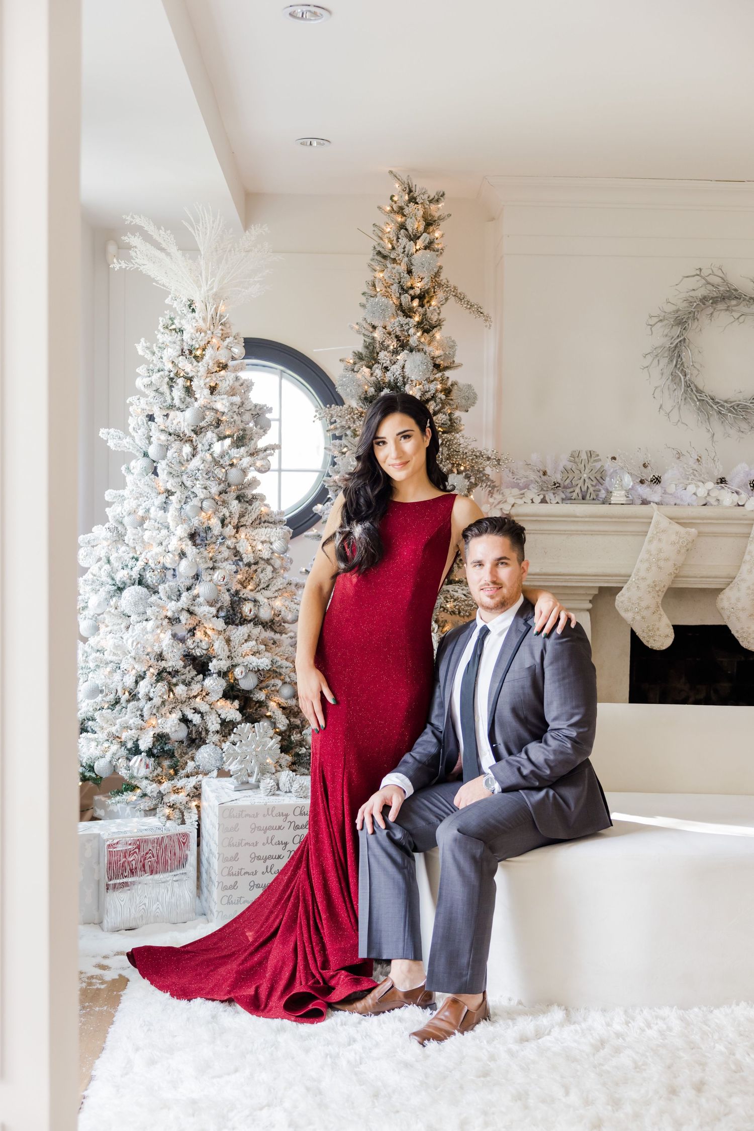 Glamorous holiday portrait with flowing red gown against white Christmas trees and fireplace.