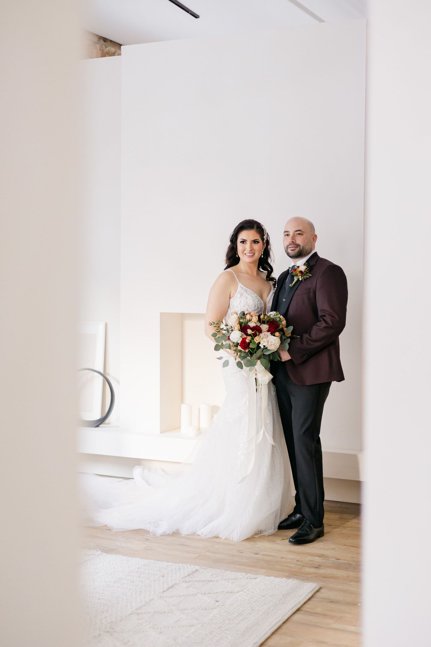 Wedding portrait framed through doorway showing couple in burgundy suit and white lace gown.