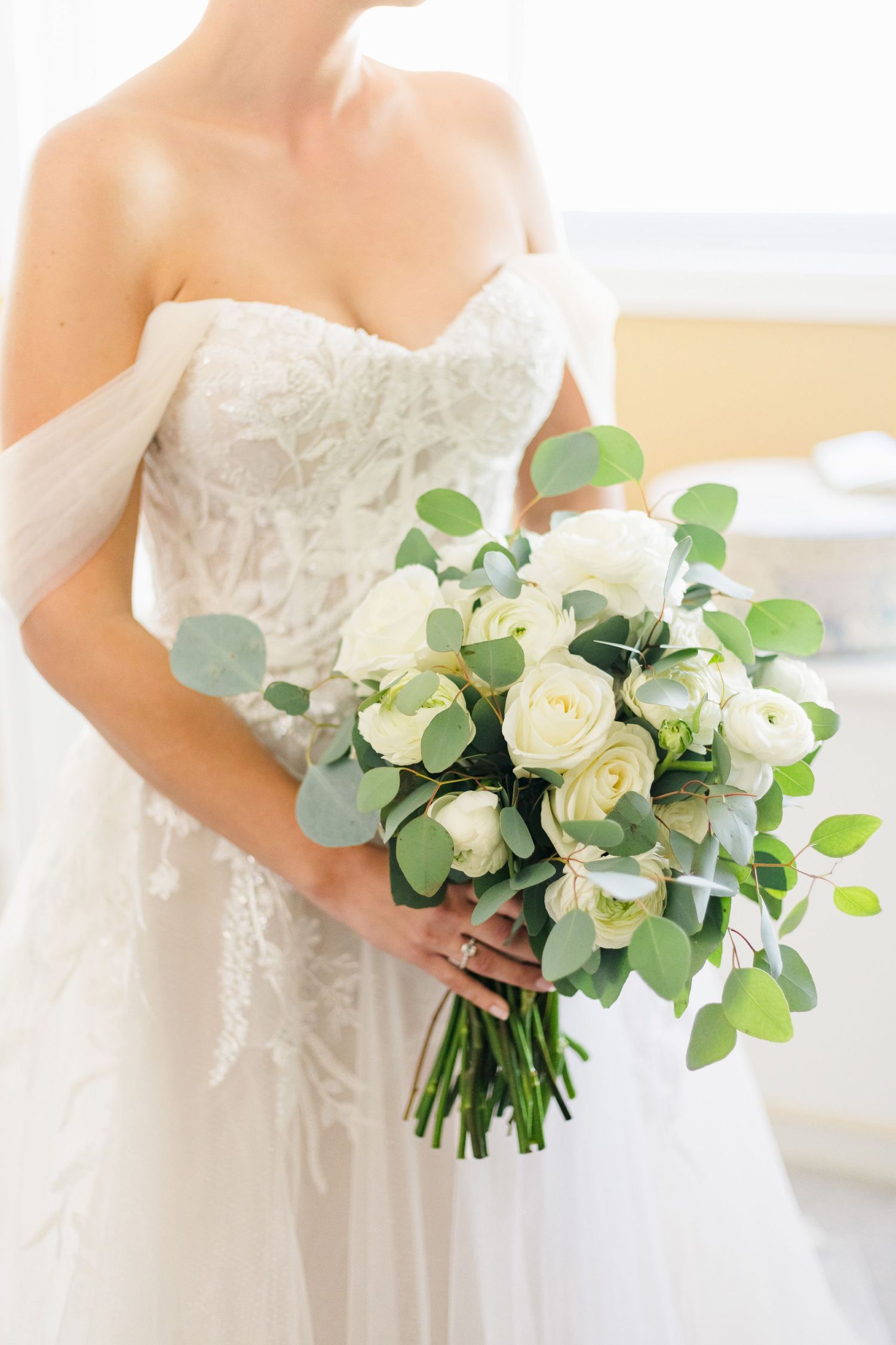 Elegant white bridal bouquet with roses and eucalyptus leaves against lace wedding dress.