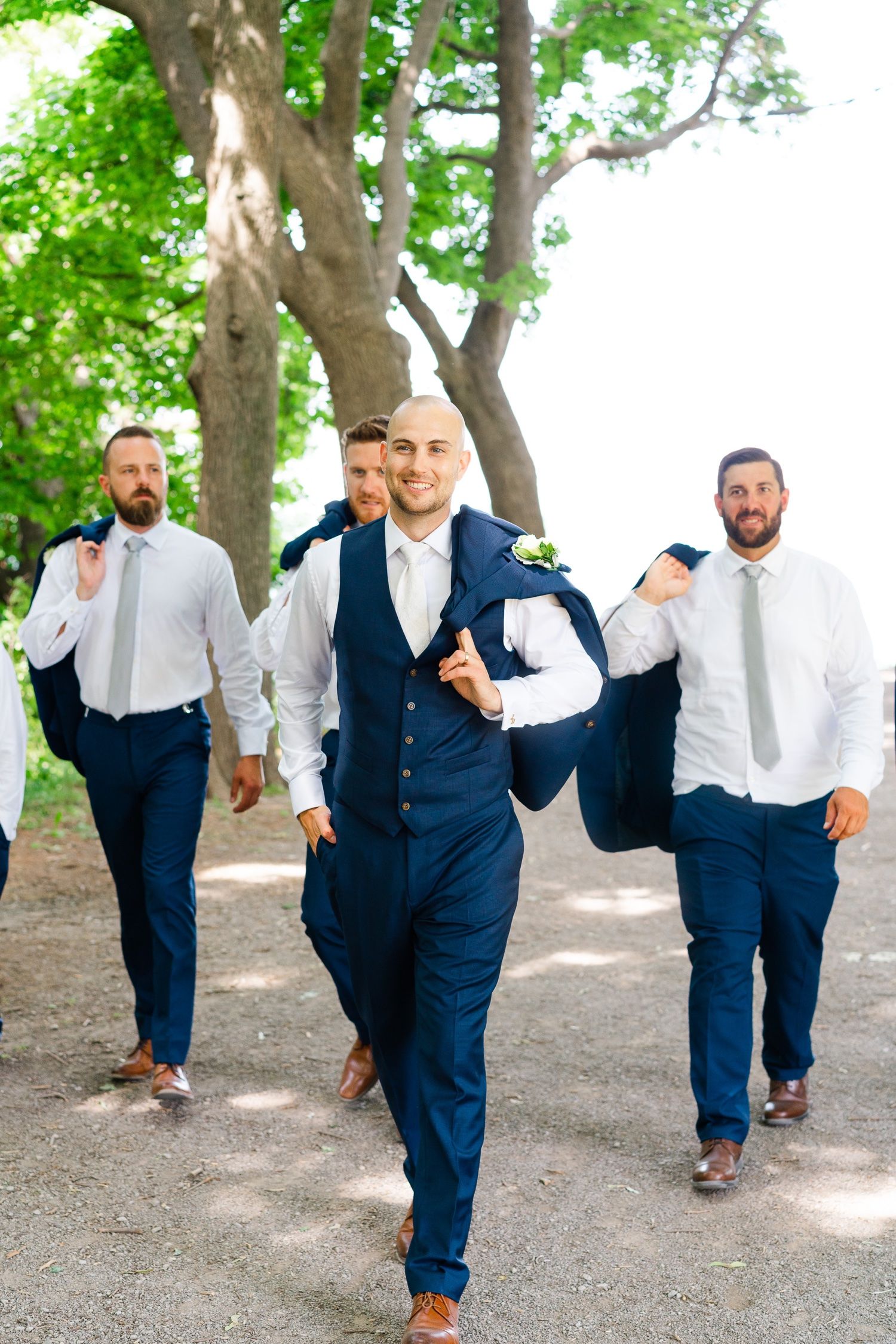 Groomsmen in navy suits and white shirts walk playfully down a tree-lined path.