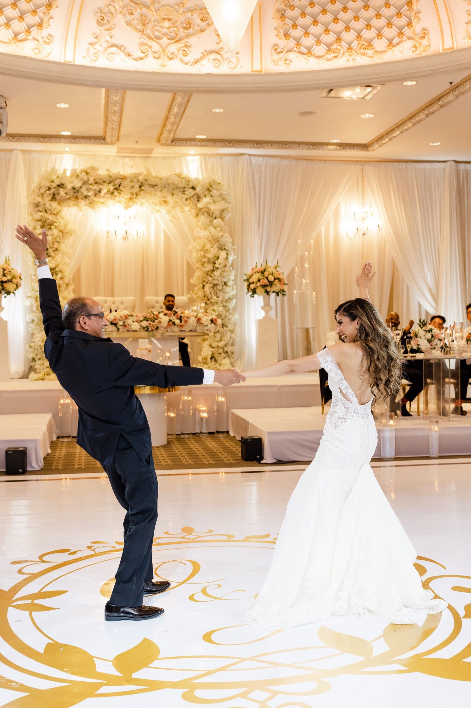 Dancing couple spins on an ornate gold and white dance floor beneath a floral archway at their wedding reception.