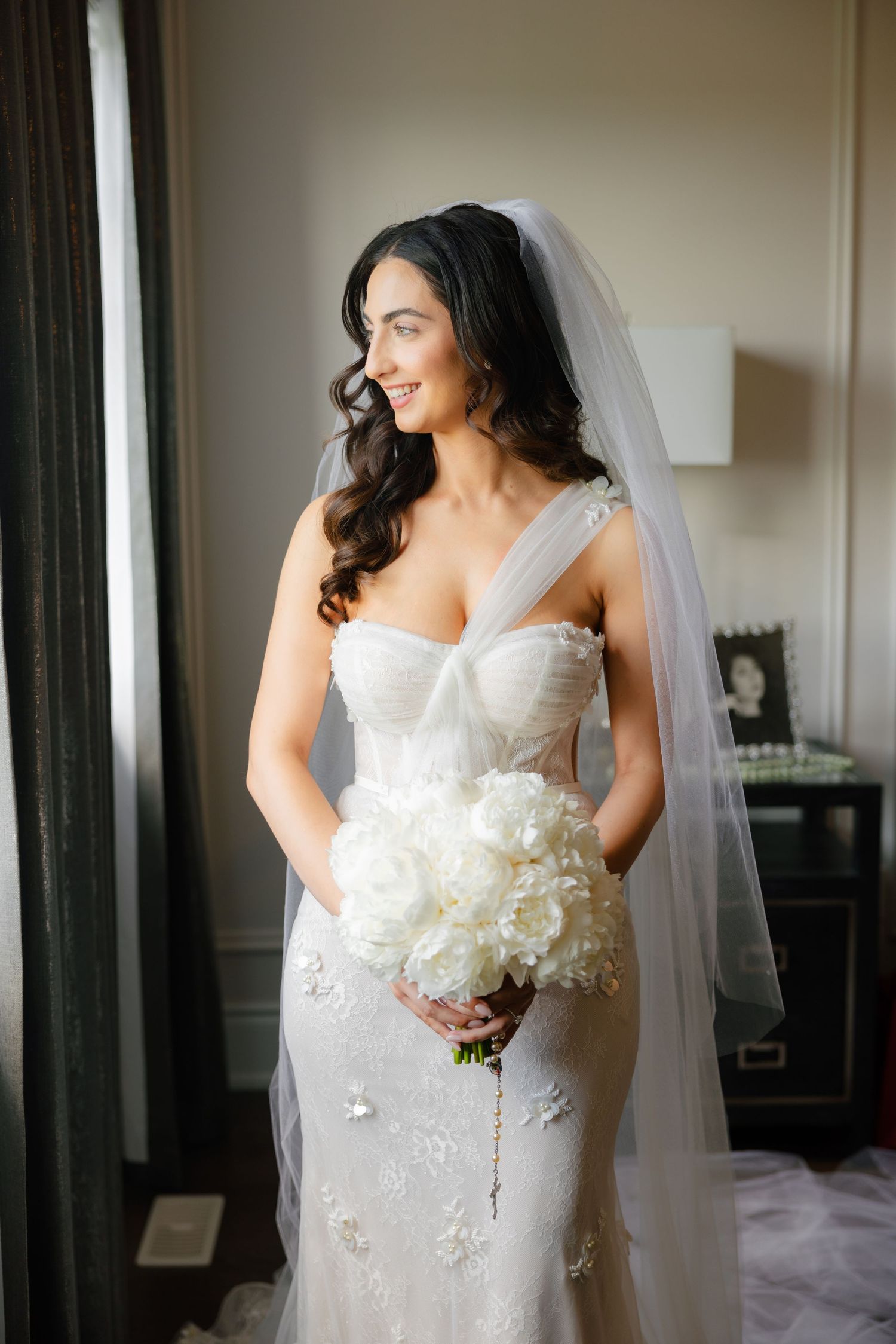 Elegant bride poses by window holding a luxurious white peony bouquet.