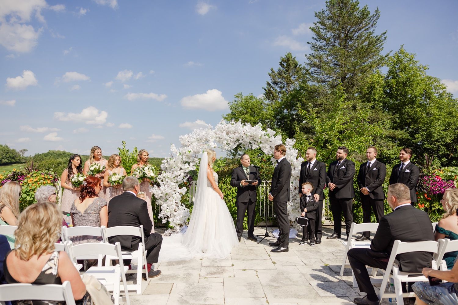 An outdoor wedding ceremony setup with white floral arch and chairs on a stone terrace.