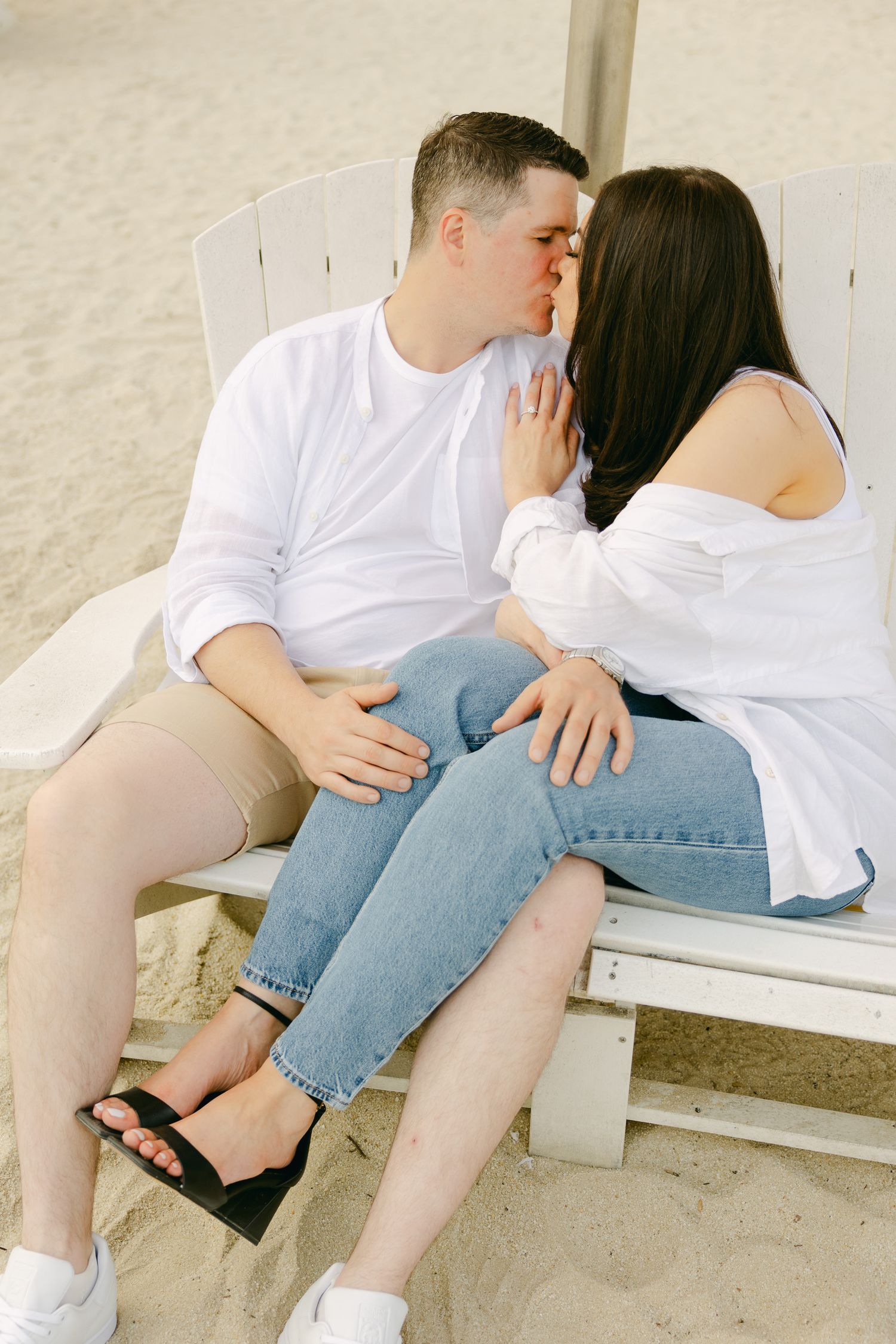 Romantic couple in casual white outfits embracing on white wooden bench.