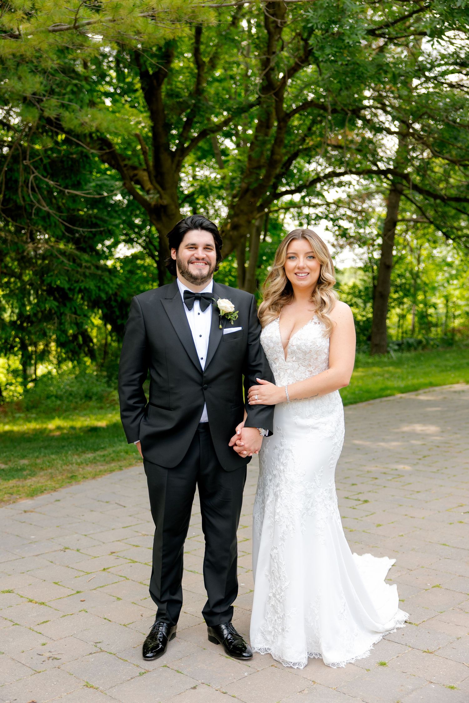 Bride in lace gown and groom in tuxedo pose together on garden pathway.