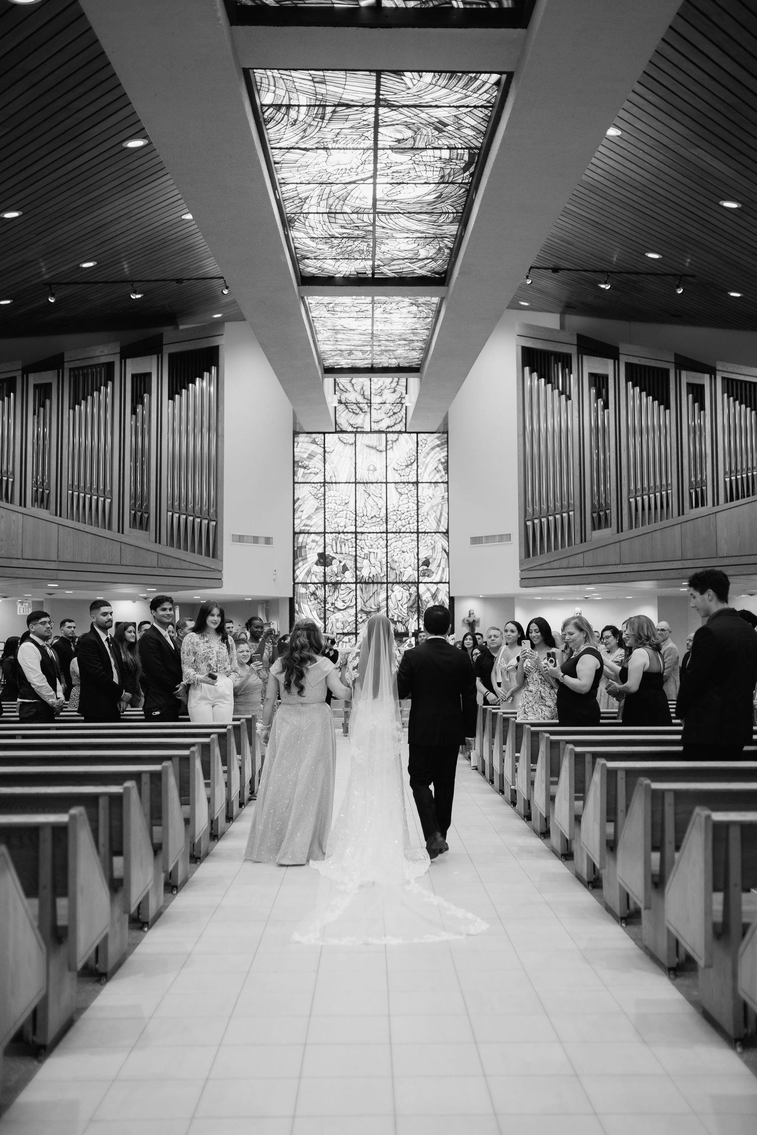 Black and white photo of wedding ceremony in modern church with pipe organs and glass ceiling.