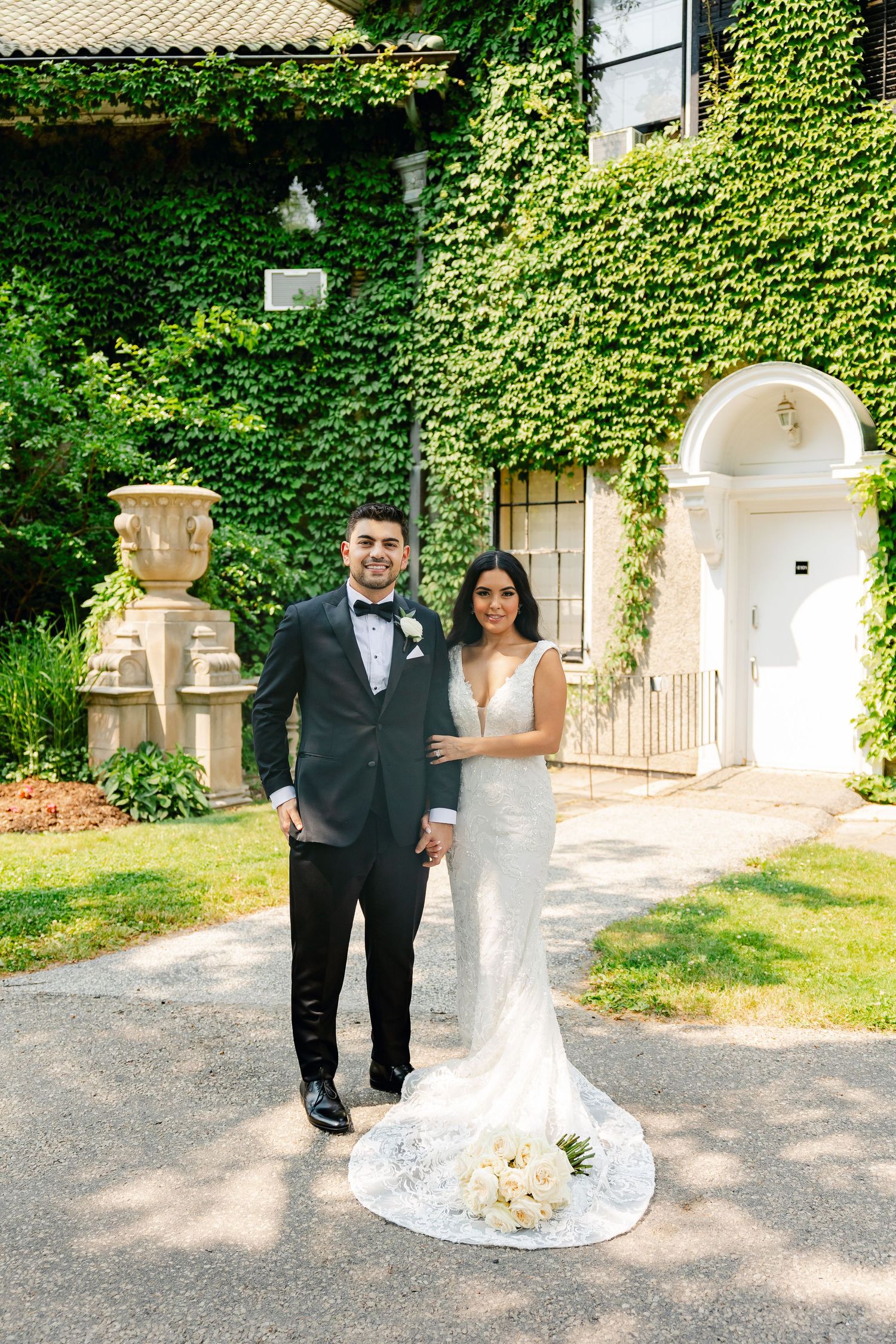 Wedding couple posing by antique urn and ivy-covered wall in formal attire.