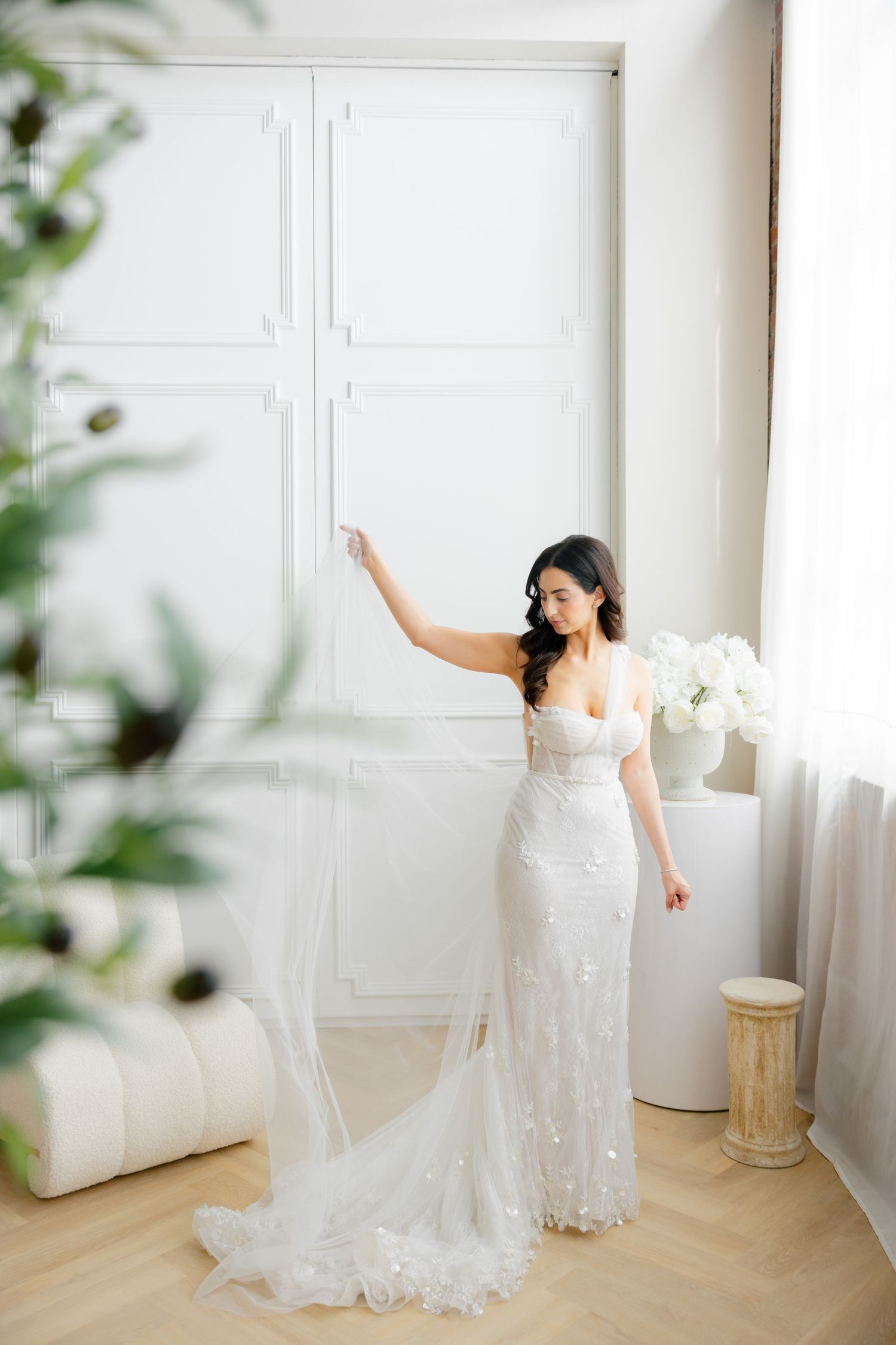 Bride in fitted lace gown poses elegantly by white paneled wall with trailing veil.