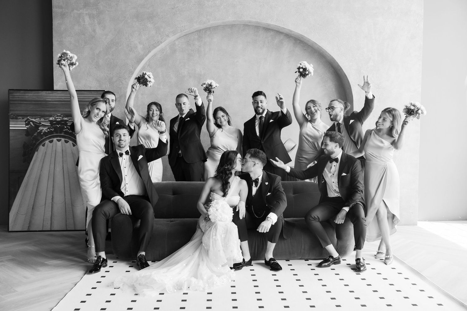 Wedding party strikes fun poses against gray archway in black and white photo.