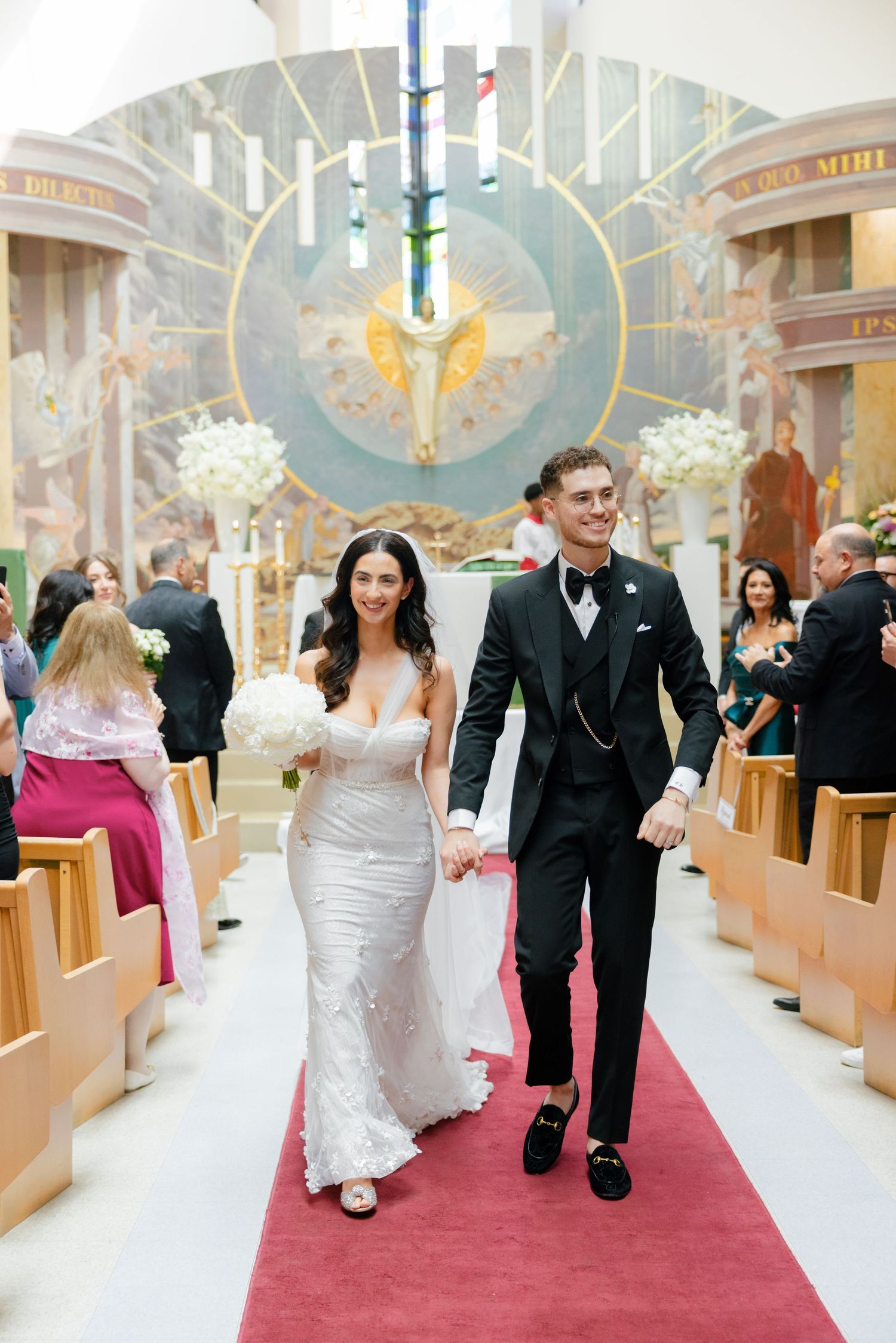 A newly married couple walks down the aisle of an ornate church with a red carpet and decorative crucifix.