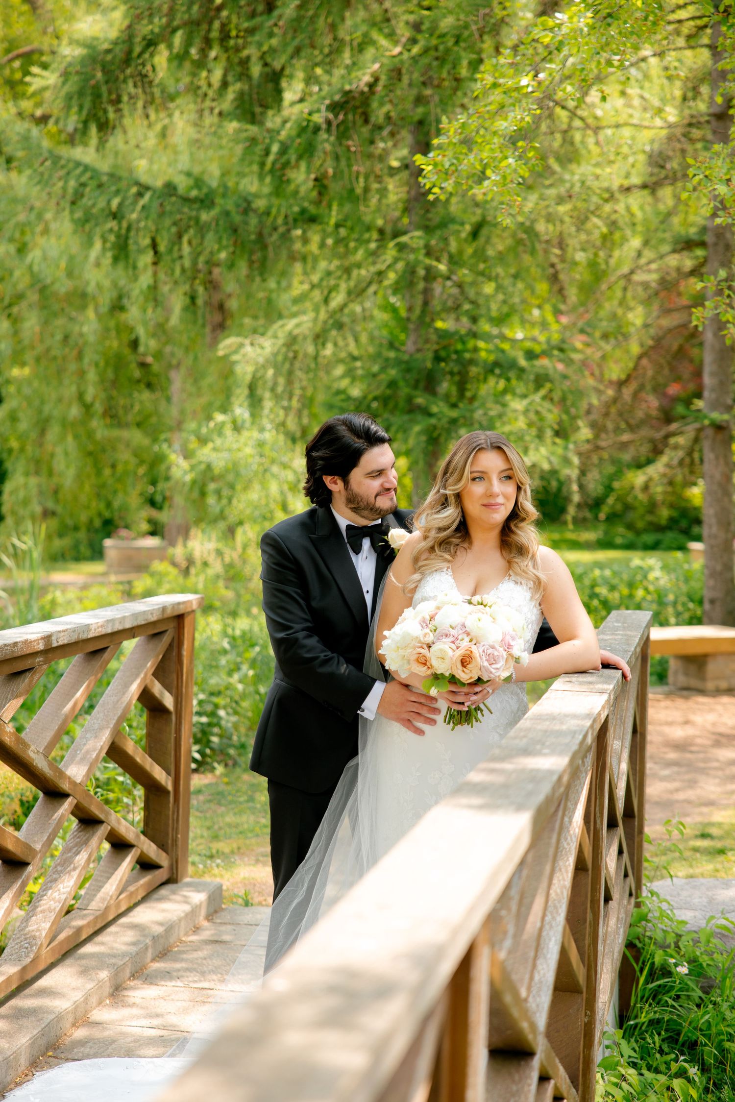 Romantic wedding moment captured on rustic bridge with blush bouquet and trailing veil.