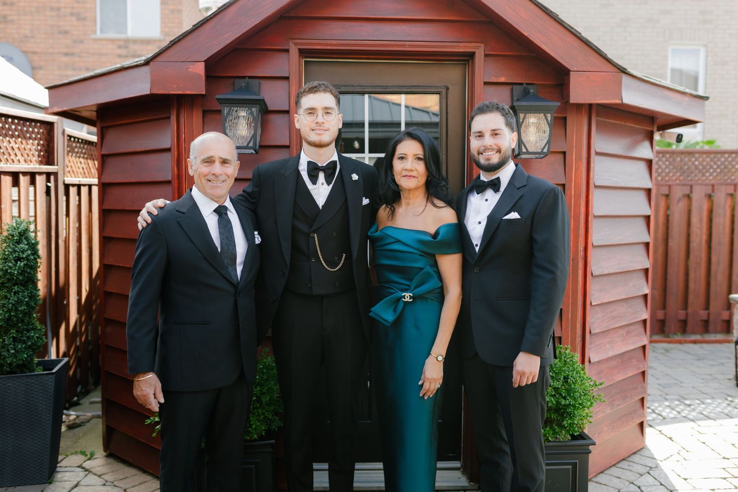 A group poses in formal attire against a red wooden shed backdrop.