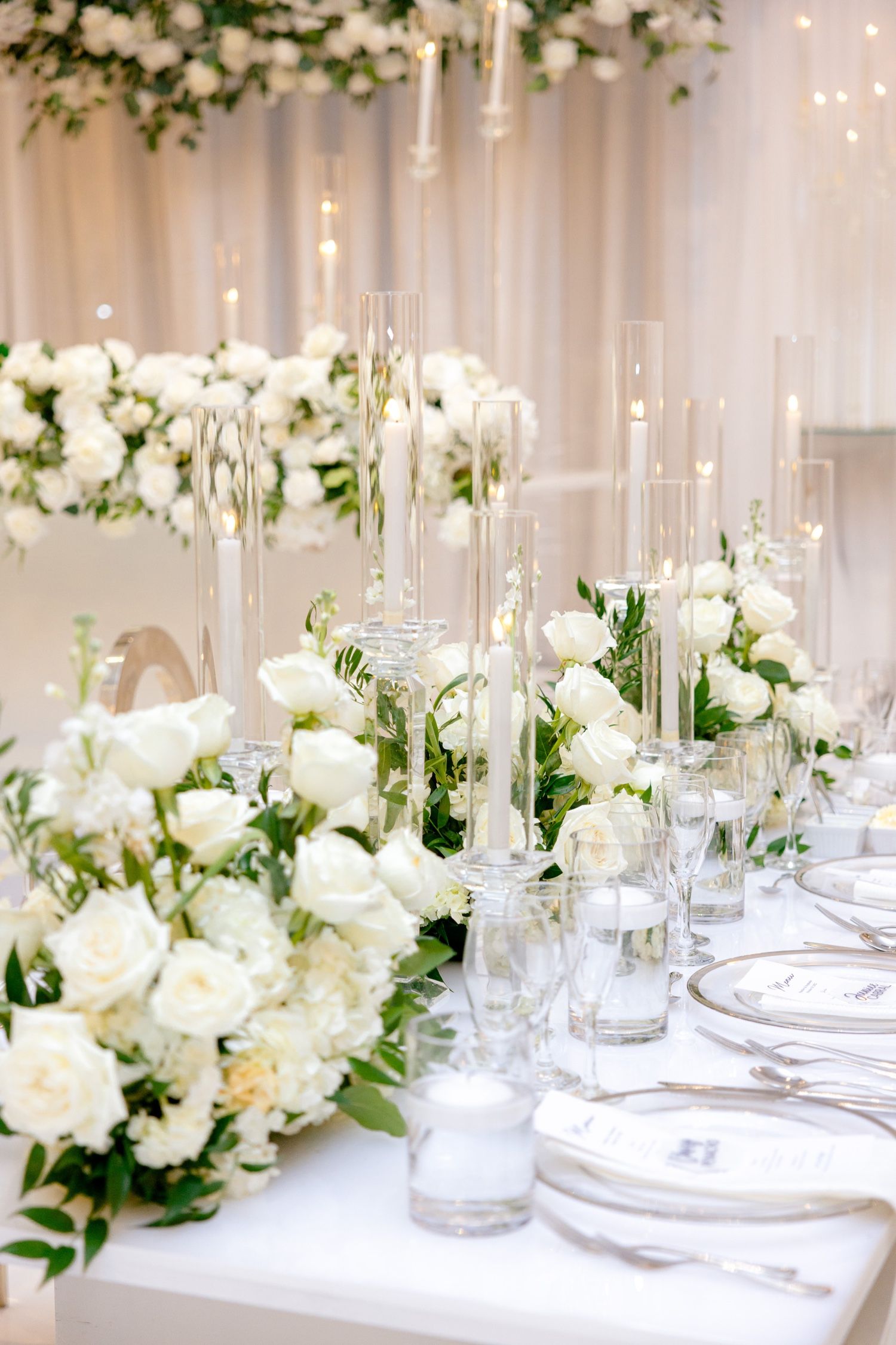 Luxurious white rose centerpieces with tall glass candleholders on an elegantly set wedding reception table.