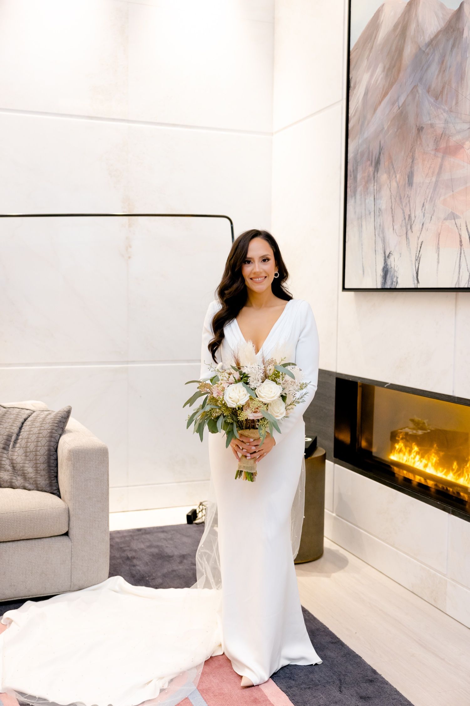 Elegant bride in flowing white gown holds beautiful bouquet of white flowers in modern room with fireplace.