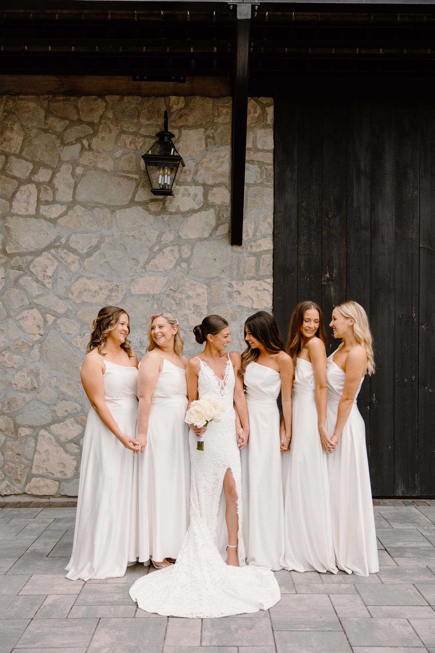 Elegant bridal party in matching white gowns gathered for portrait against rustic stone backdrop.