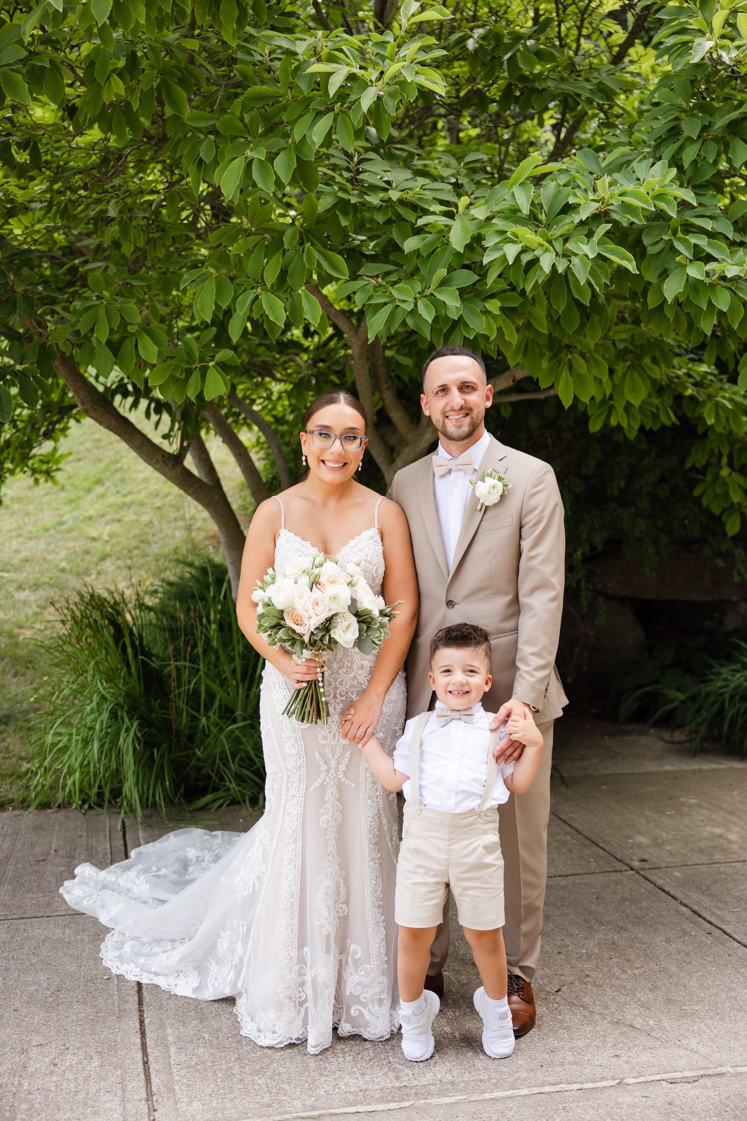 Wedding portrait of couple and young ring bearer under leafy tree.