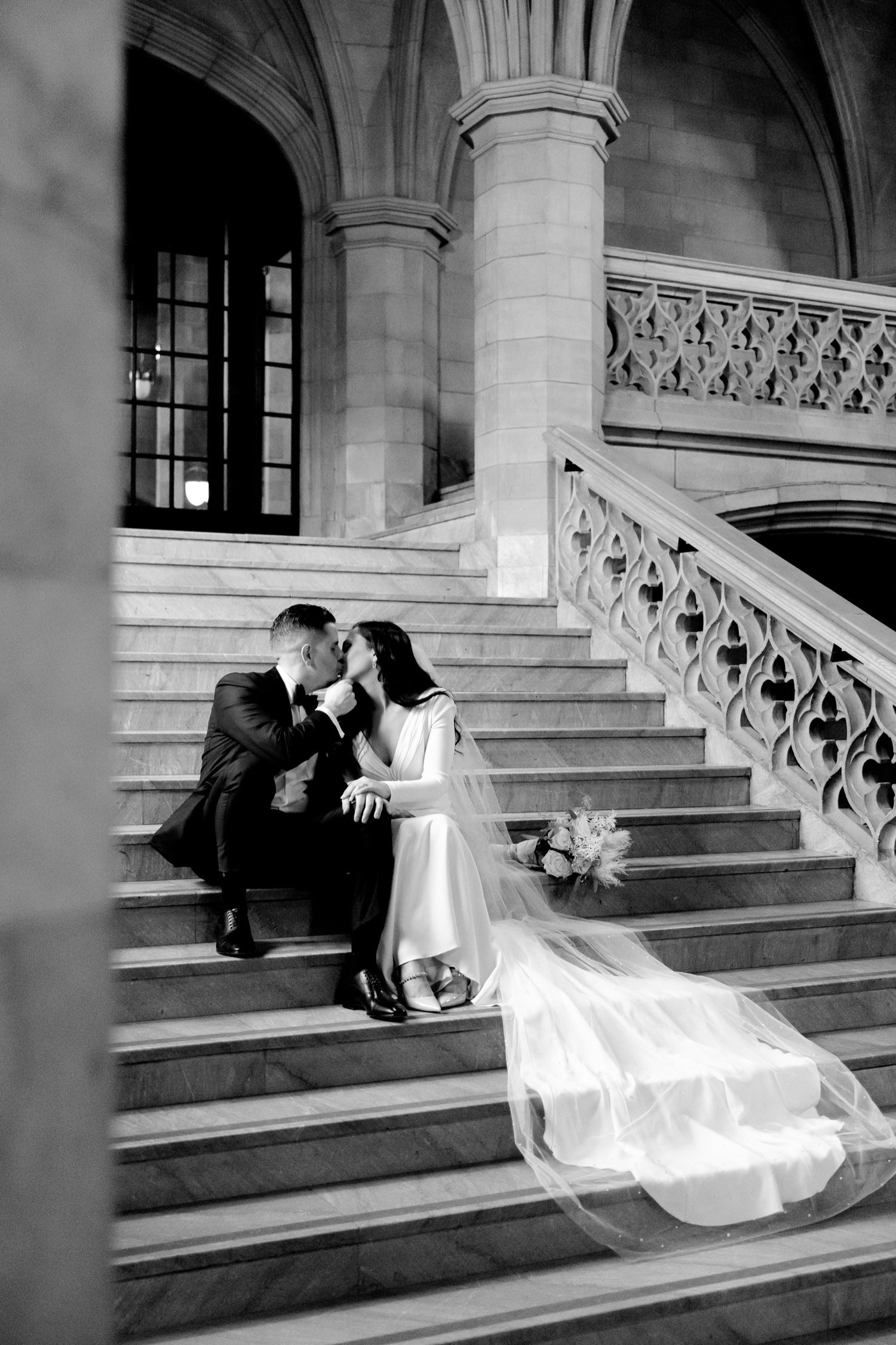 A romantic black and white wedding photo of a couple sitting on grand stone steps with flowing veil.