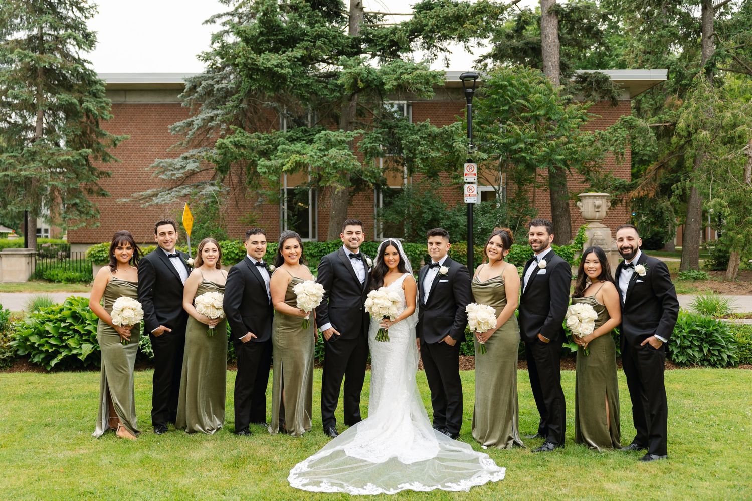 Complete wedding party lined up for a formal photo in front of a brick building with evergreen trees.