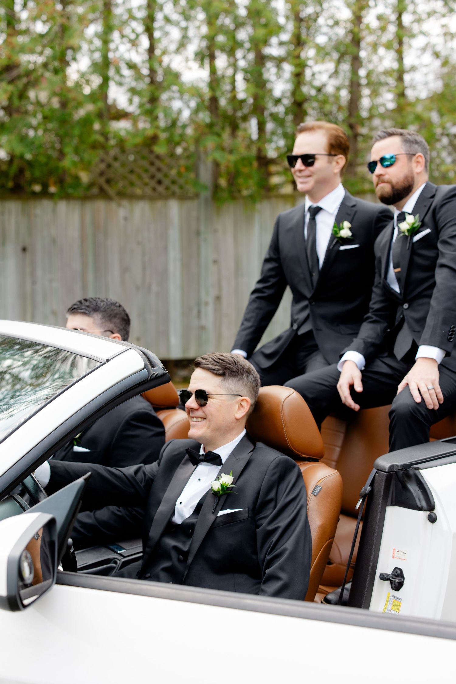 Four people in dark suits and sunglasses arrange themselves around a white luxury convertible.
