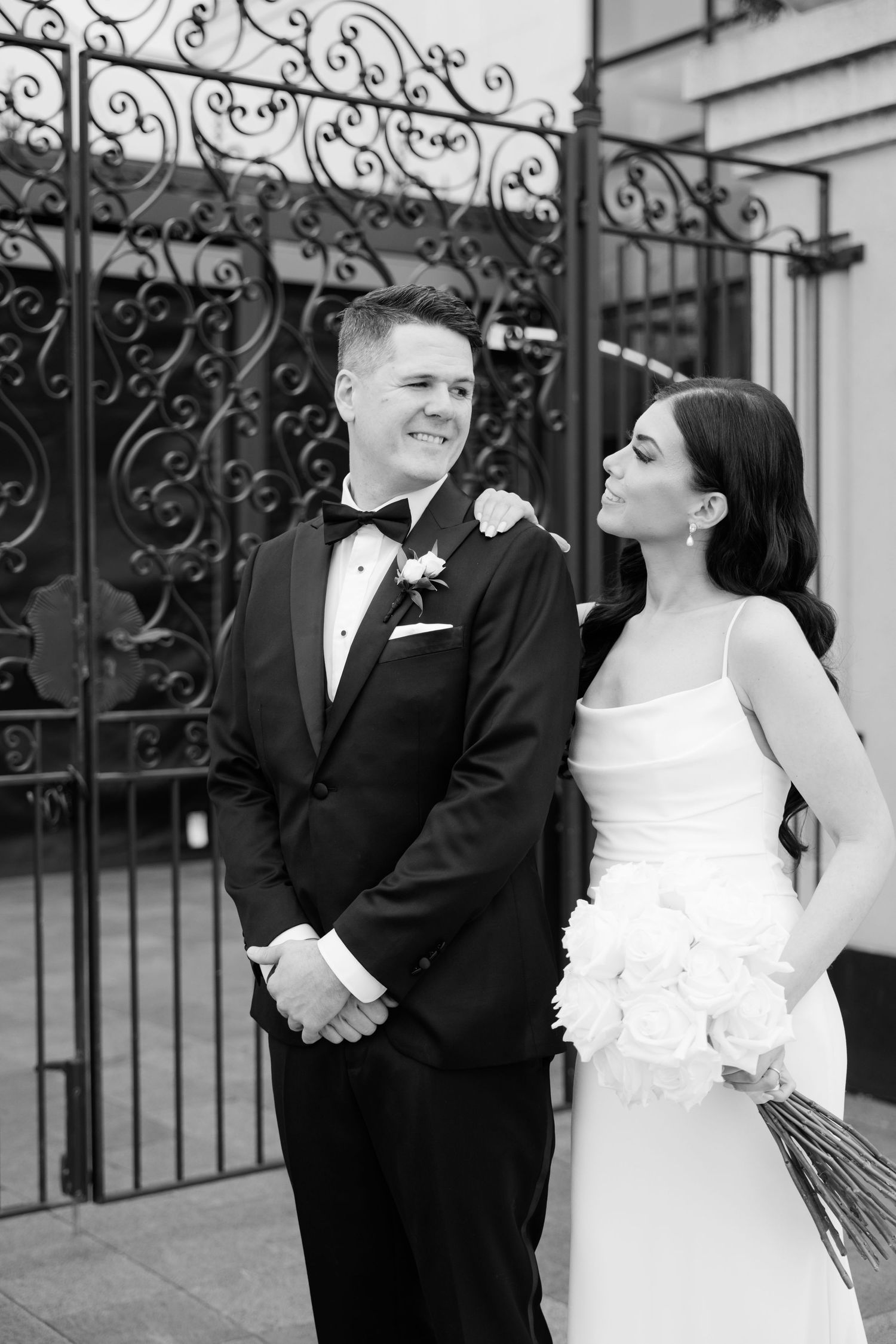 Black and white portrait of newlyweds smiling together by decorative iron gates.