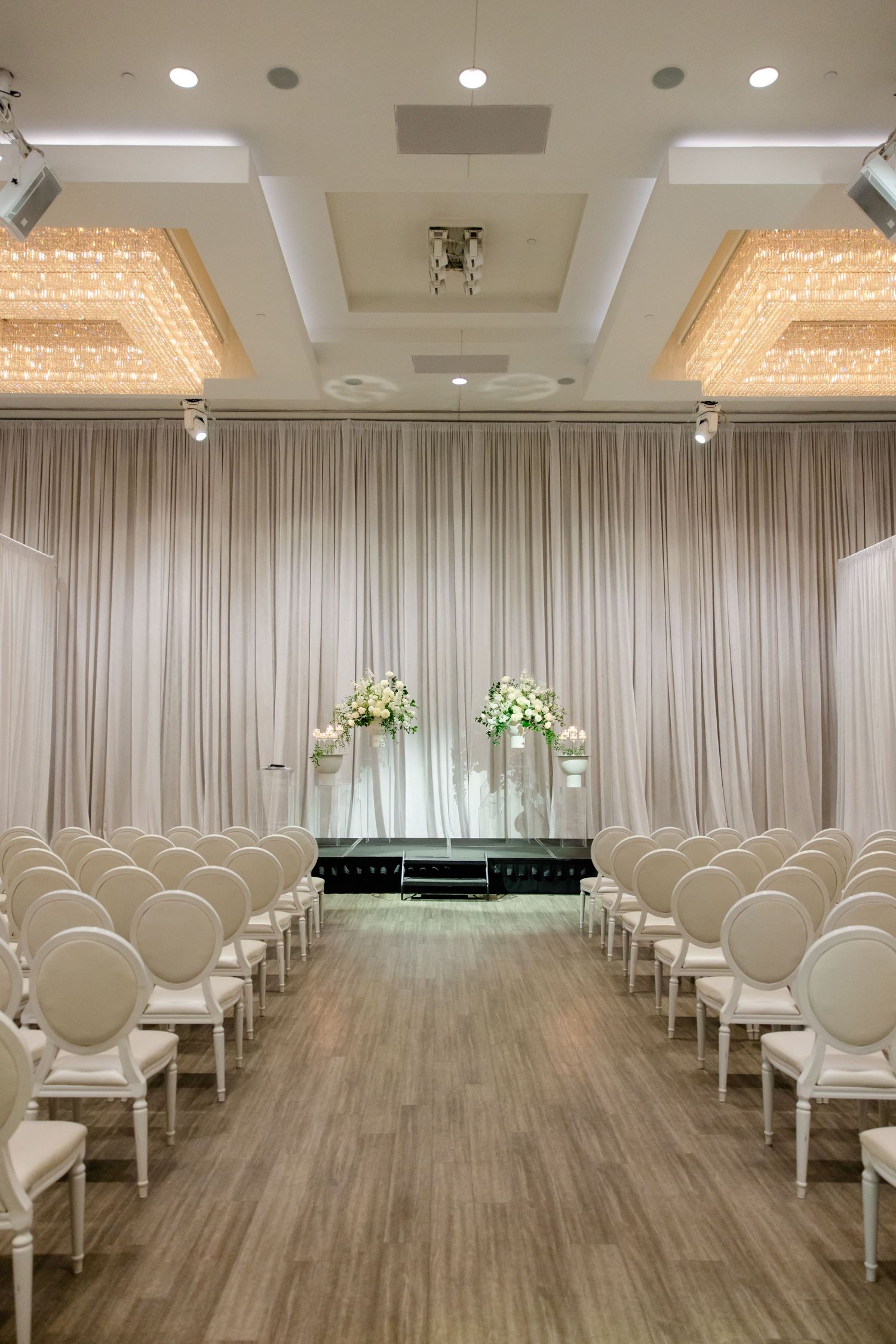 Elegant wedding ceremony venue with white drapes, crystal chandeliers and white chairs in neat rows.