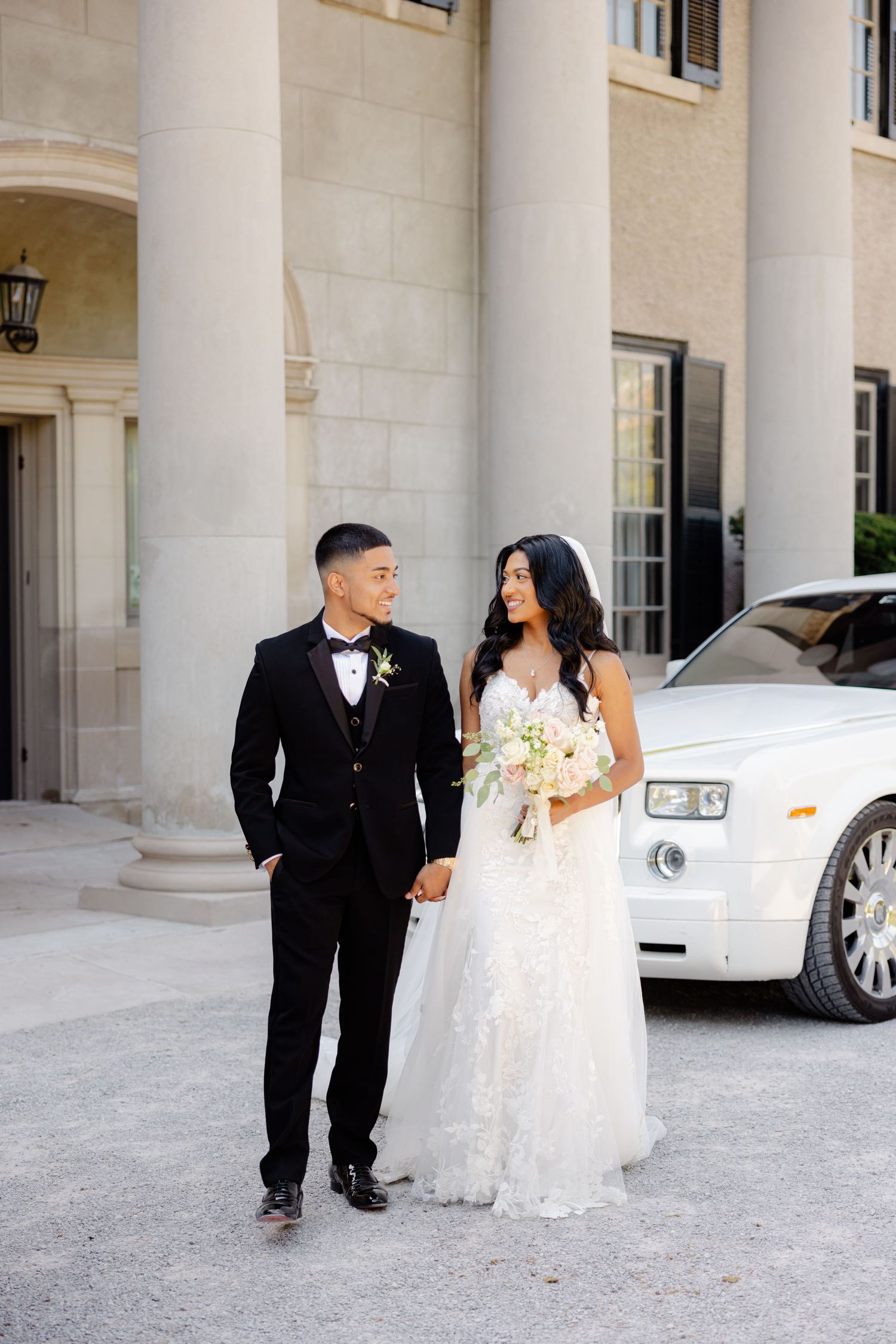 Wedding couple poses in formal attire in front of a classic white Rolls-Royce and columned building.