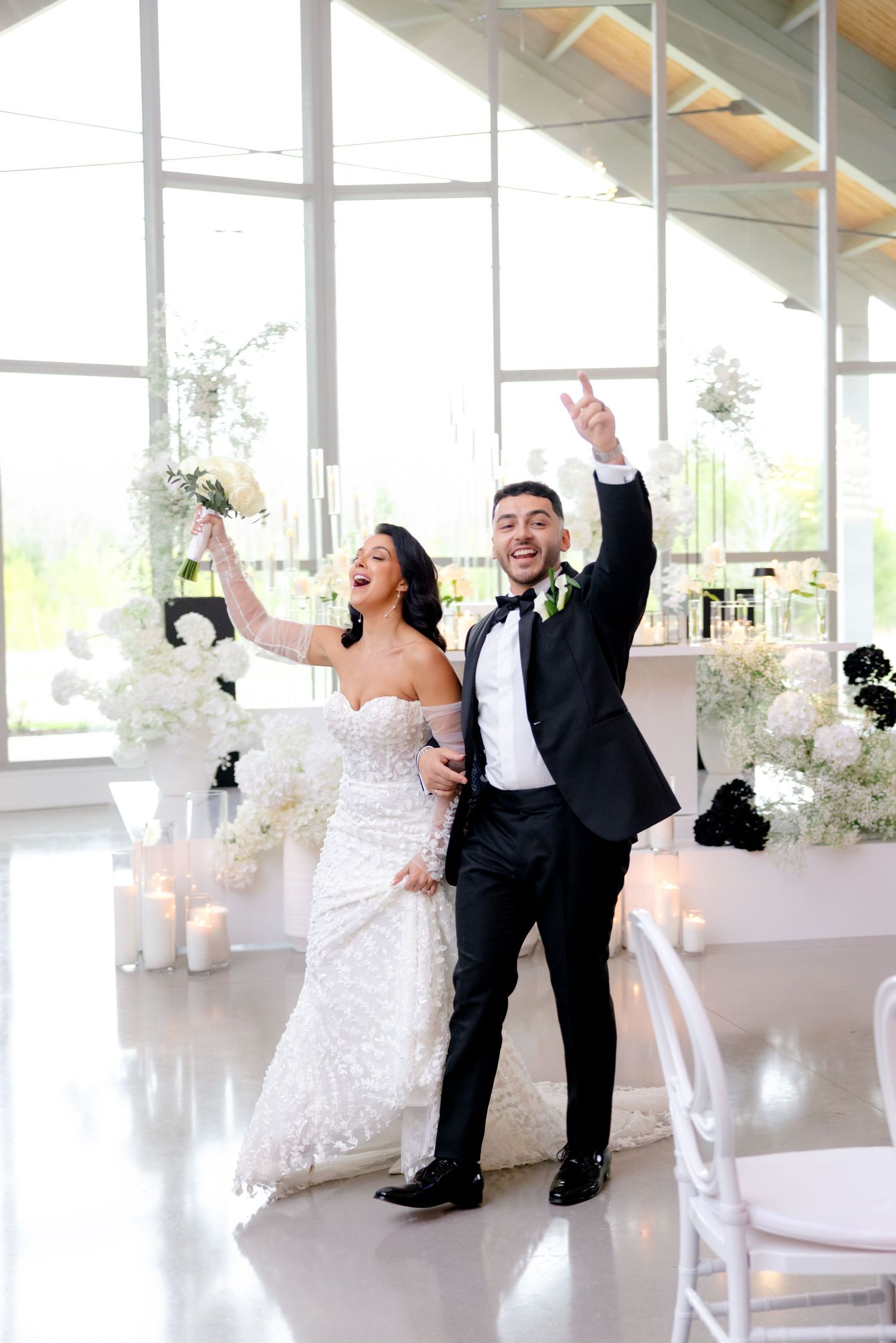 Joyful celebration moment during a wedding reception in a bright, modern venue with white floral decorations.