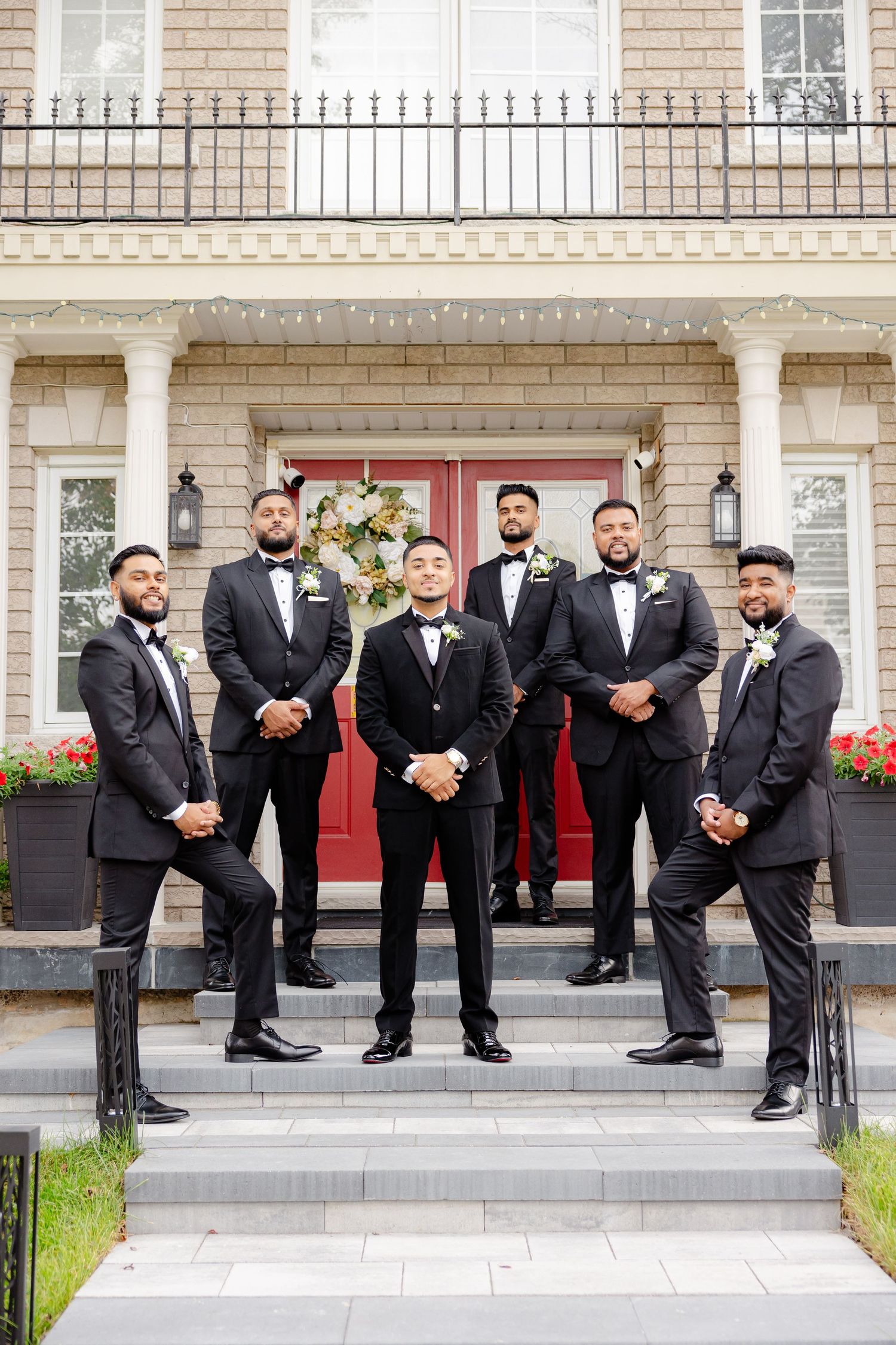 Wedding party of groomsmen in black tuxedos pose on mansion steps with red door.