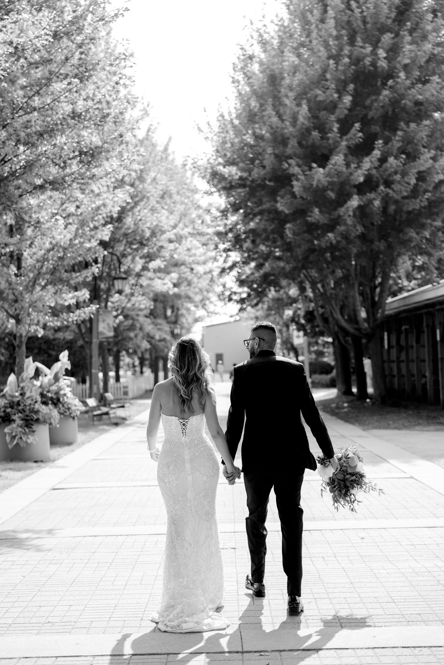 A newlywed couple walks hand in hand down a tree-lined pathway in black and white.