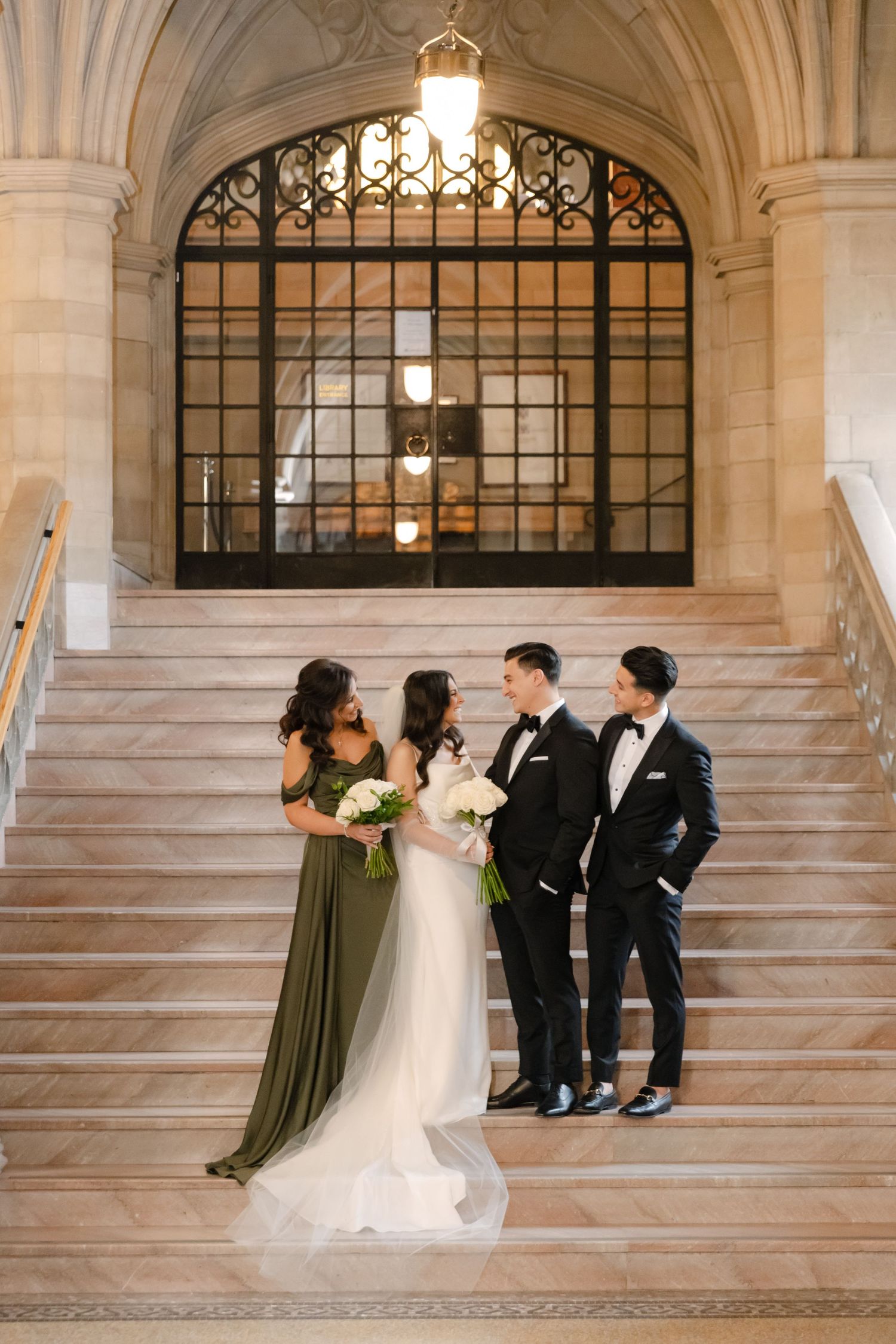 Wedding party poses on elegant marble staircase with ornate arched window and classic architecture in background.