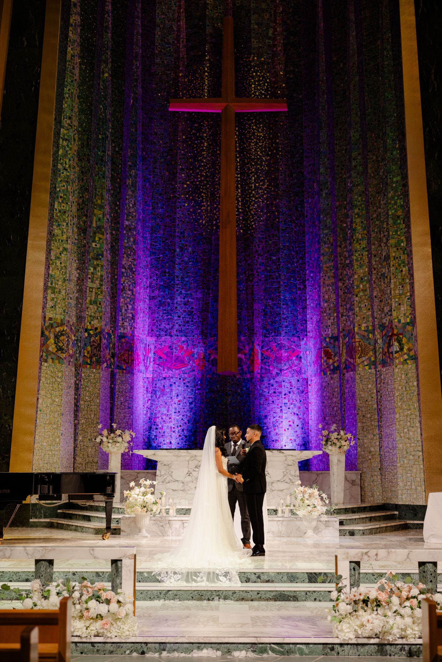 Couple stands at altar beneath illuminated cross with dramatic purple lighting in modern church setting.