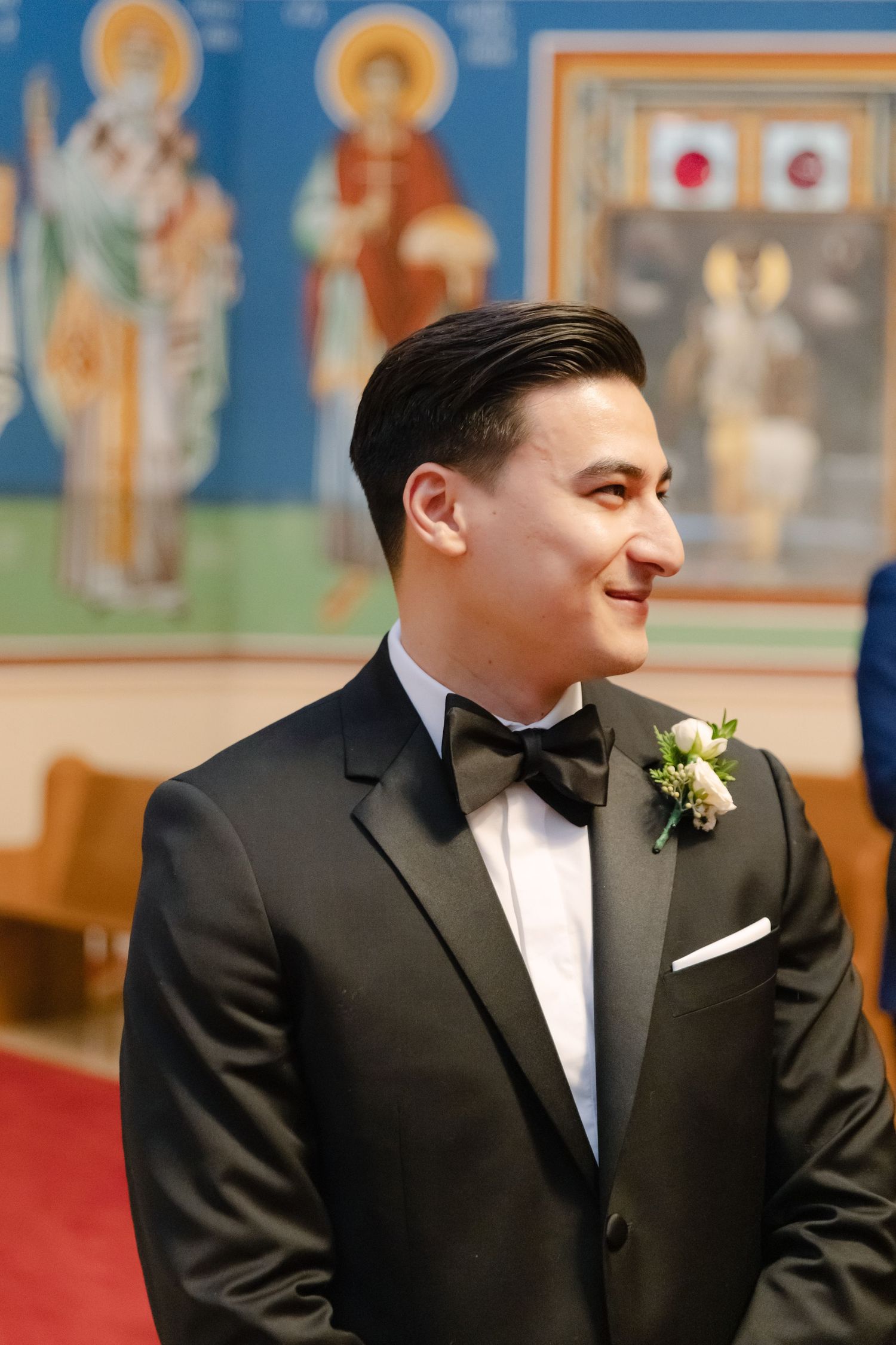 Groom in classic tuxedo stands in church with religious icons and blue walls in background.