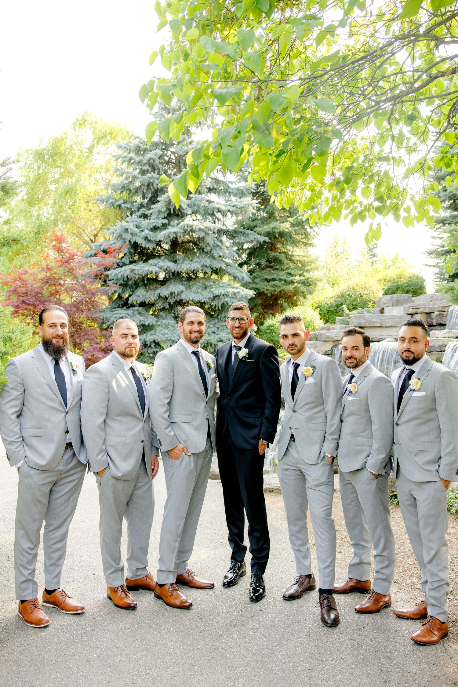 Groomsmen in light gray suits pose with groom in black suit on garden path.