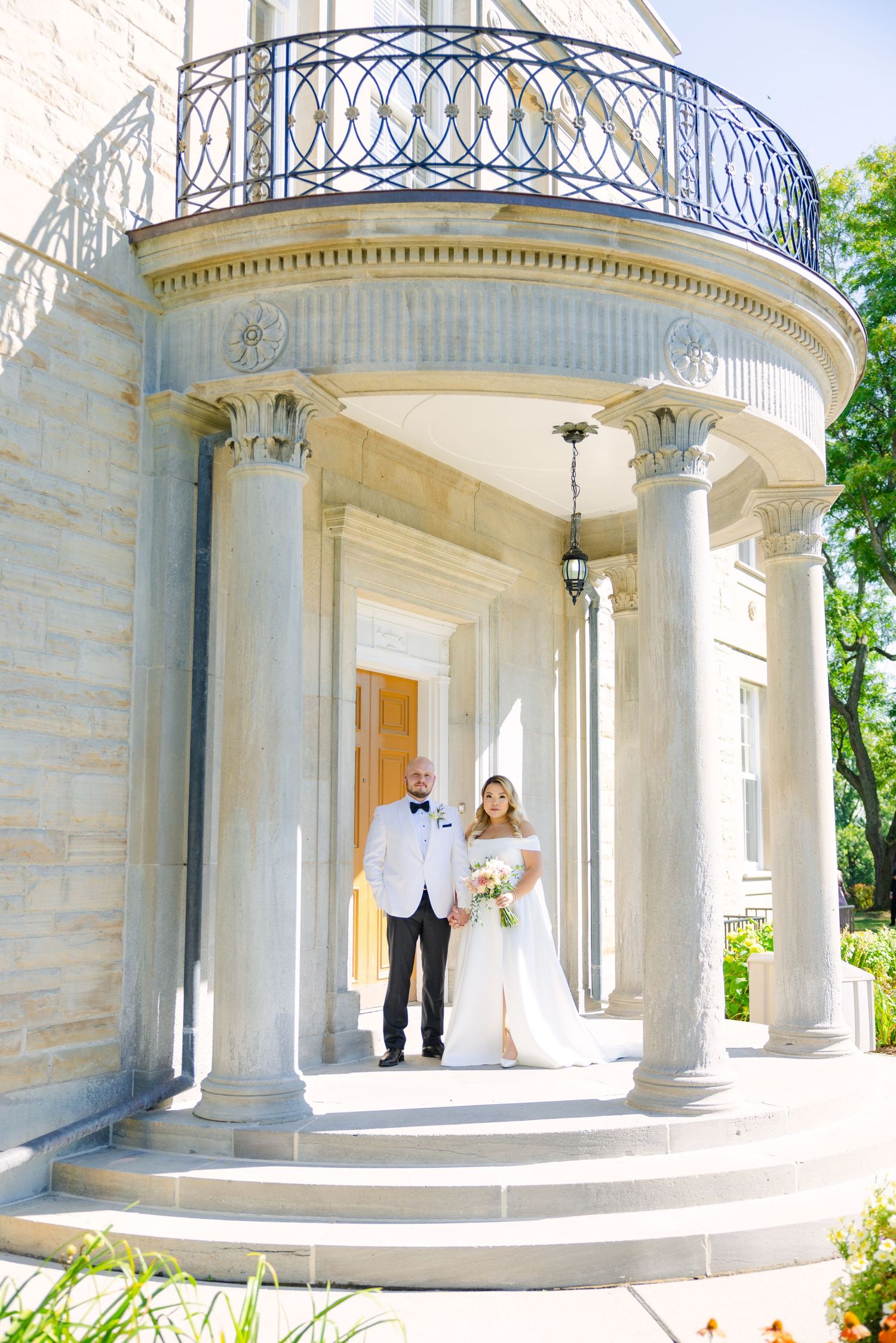 Sunlit wedding photo of a couple standing under a columned portico with wrought iron details.