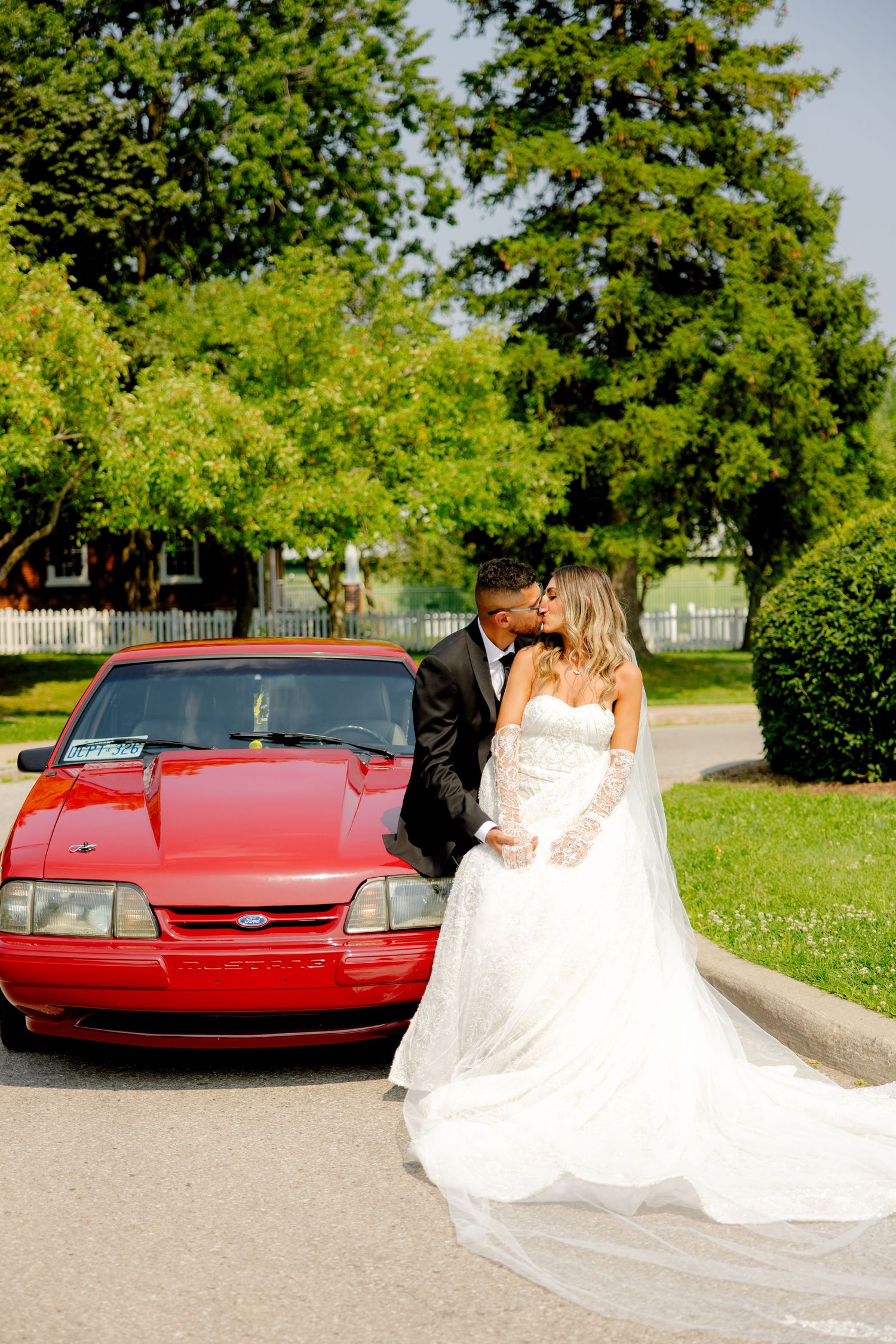 Elegant wedding couple embraces in front of a red car with a beautiful garden backdrop.
