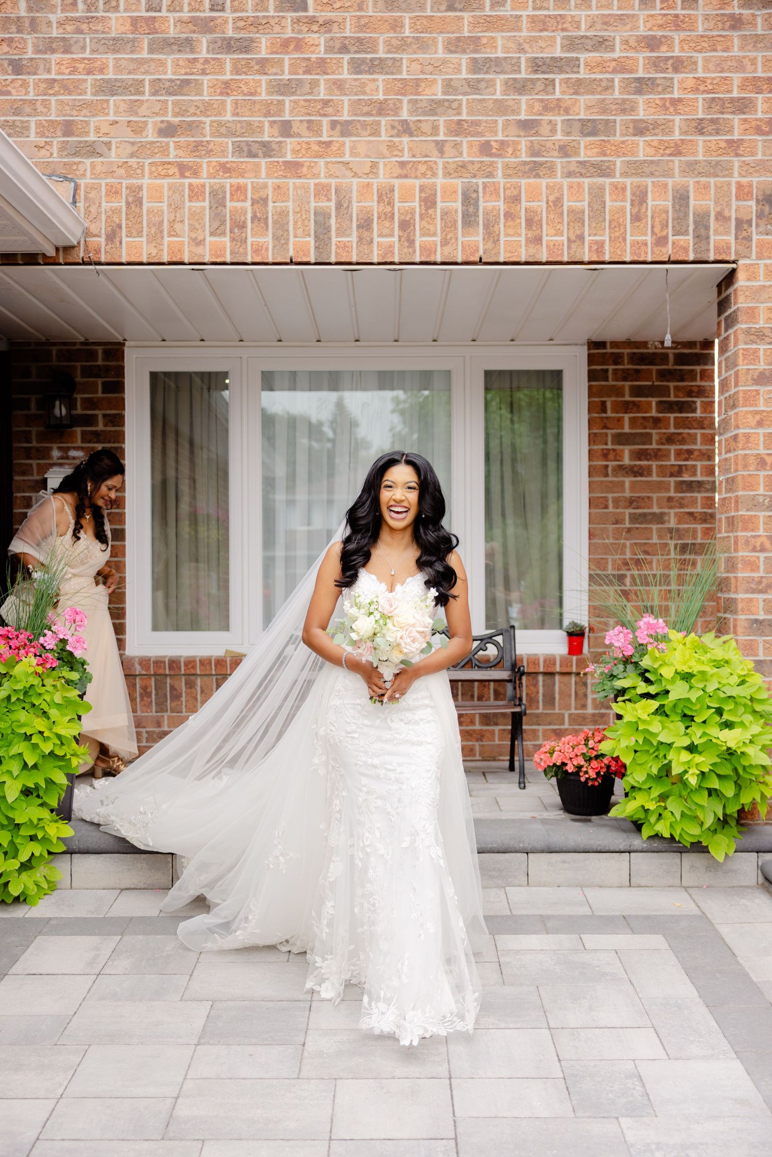 Stunning bride in a lace white wedding gown with long veil poses on front porch with brick backdrop.