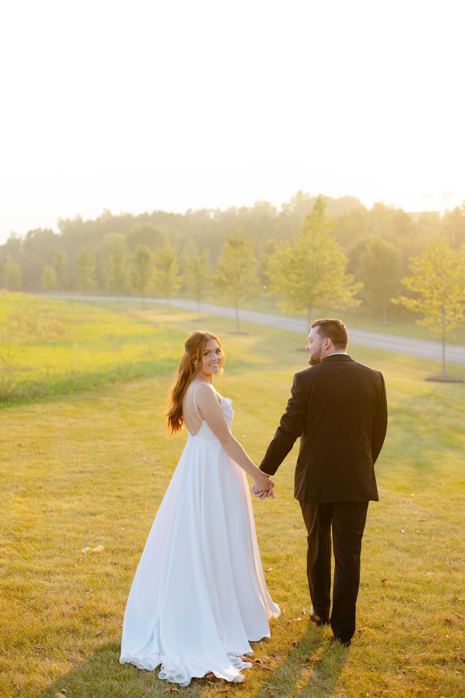 A romantic sunset wedding photo of a couple holding hands on a hill overlooking a tree-lined path.