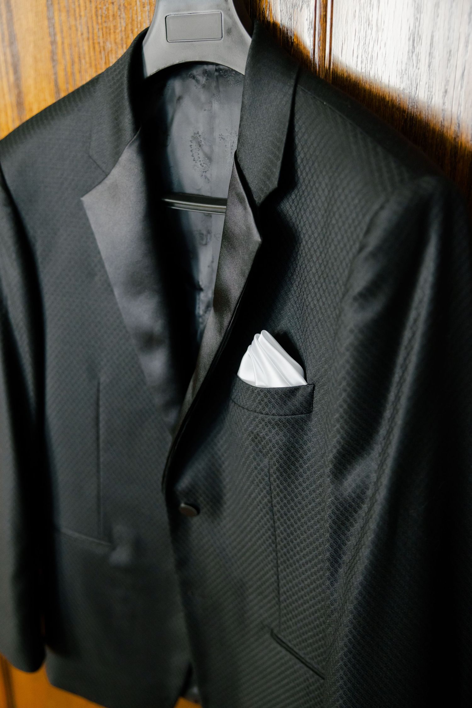Close-up of black textured tuxedo jacket with white pocket square on hanger.