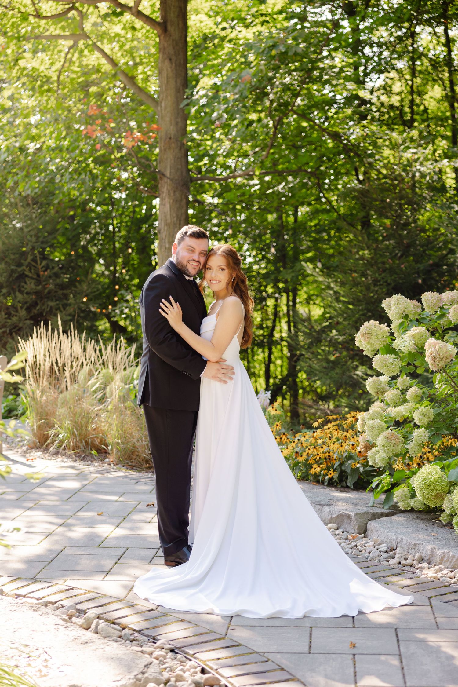 A romantic wedding couple embraces on a garden path surrounded by lush greenery and hydrangea blooms.