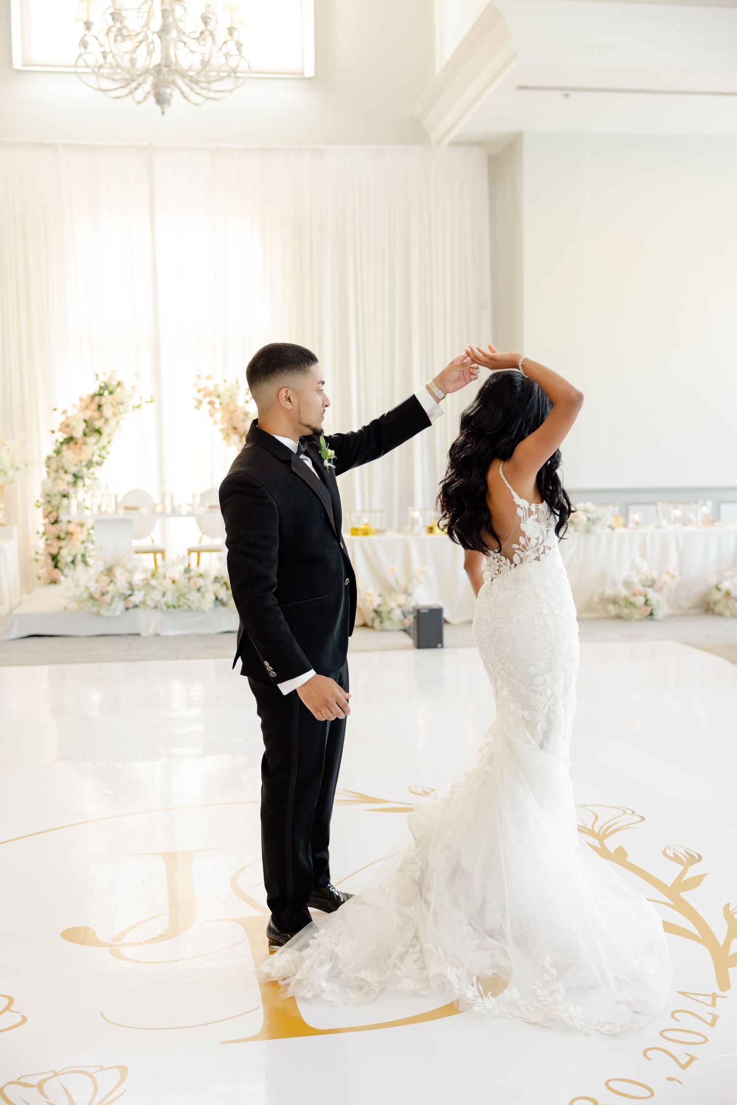 Newlyweds share a romantic dance in an elegant white ballroom with gold floor details and floral decor.