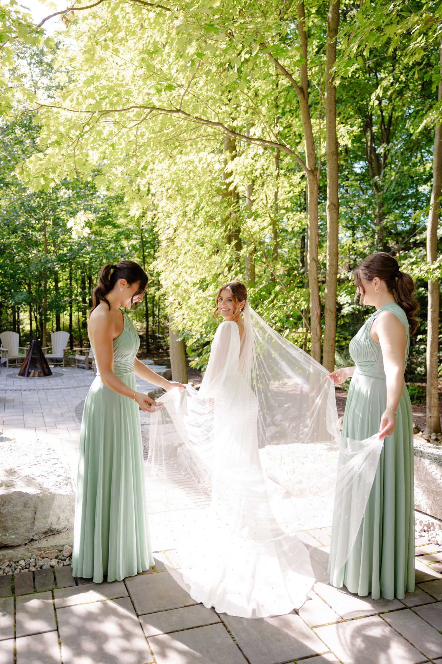 Bridesmaids in mint green gowns assist with bride's veil under sunlit tree canopy.