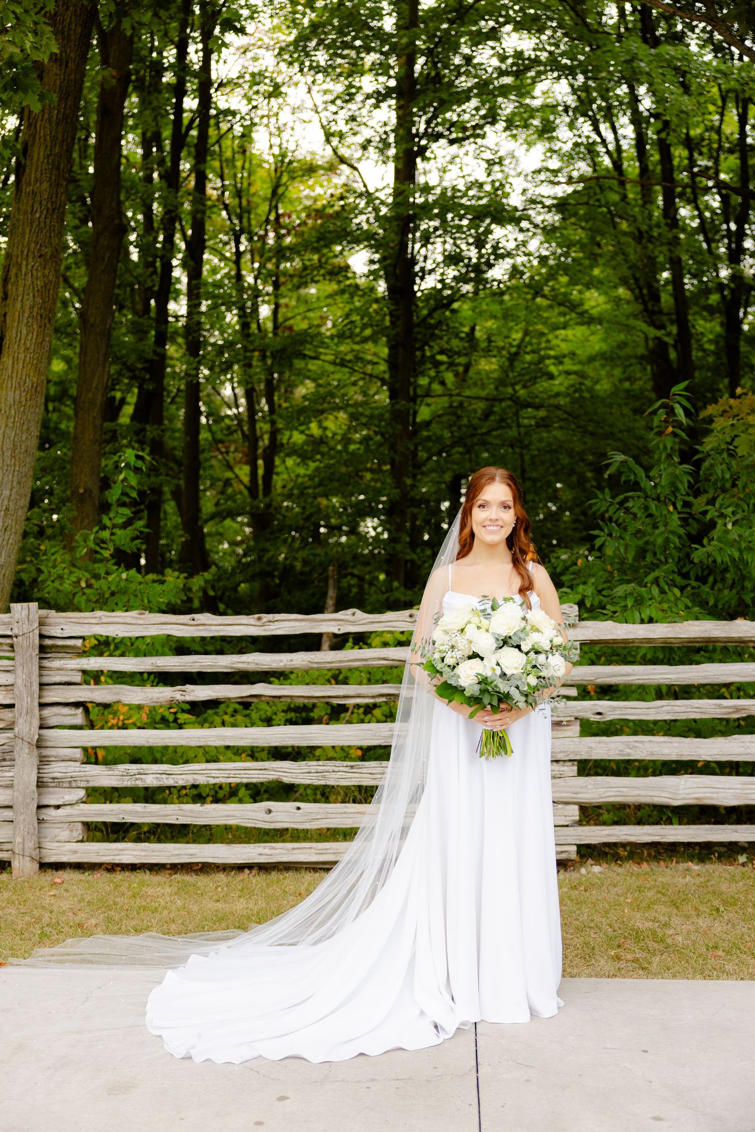 Elegant bridal portrait featuring flowing white gown and lush bouquet against rustic wooden fence.