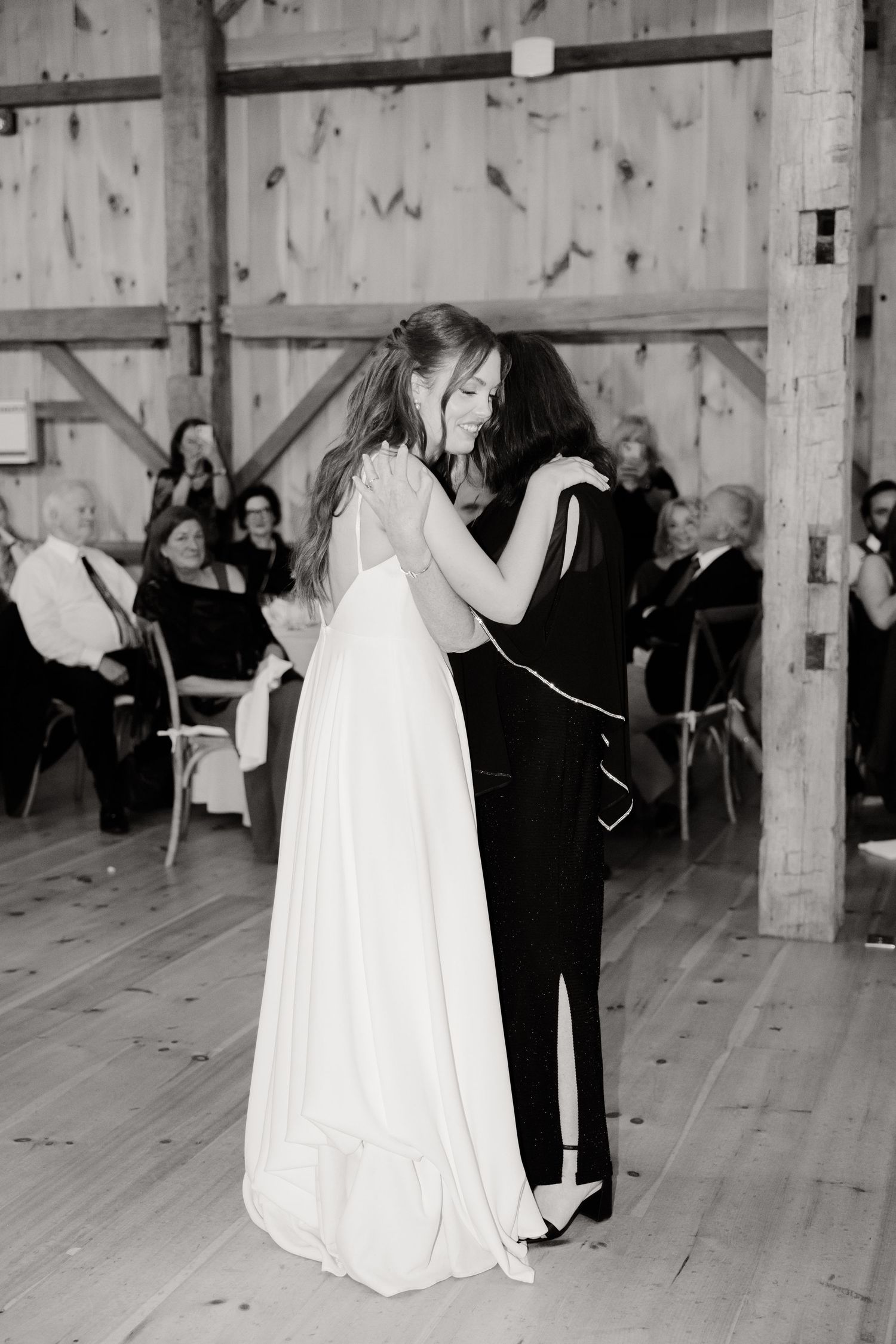A black and white photo of two people dancing at a wedding reception in a rustic barn.