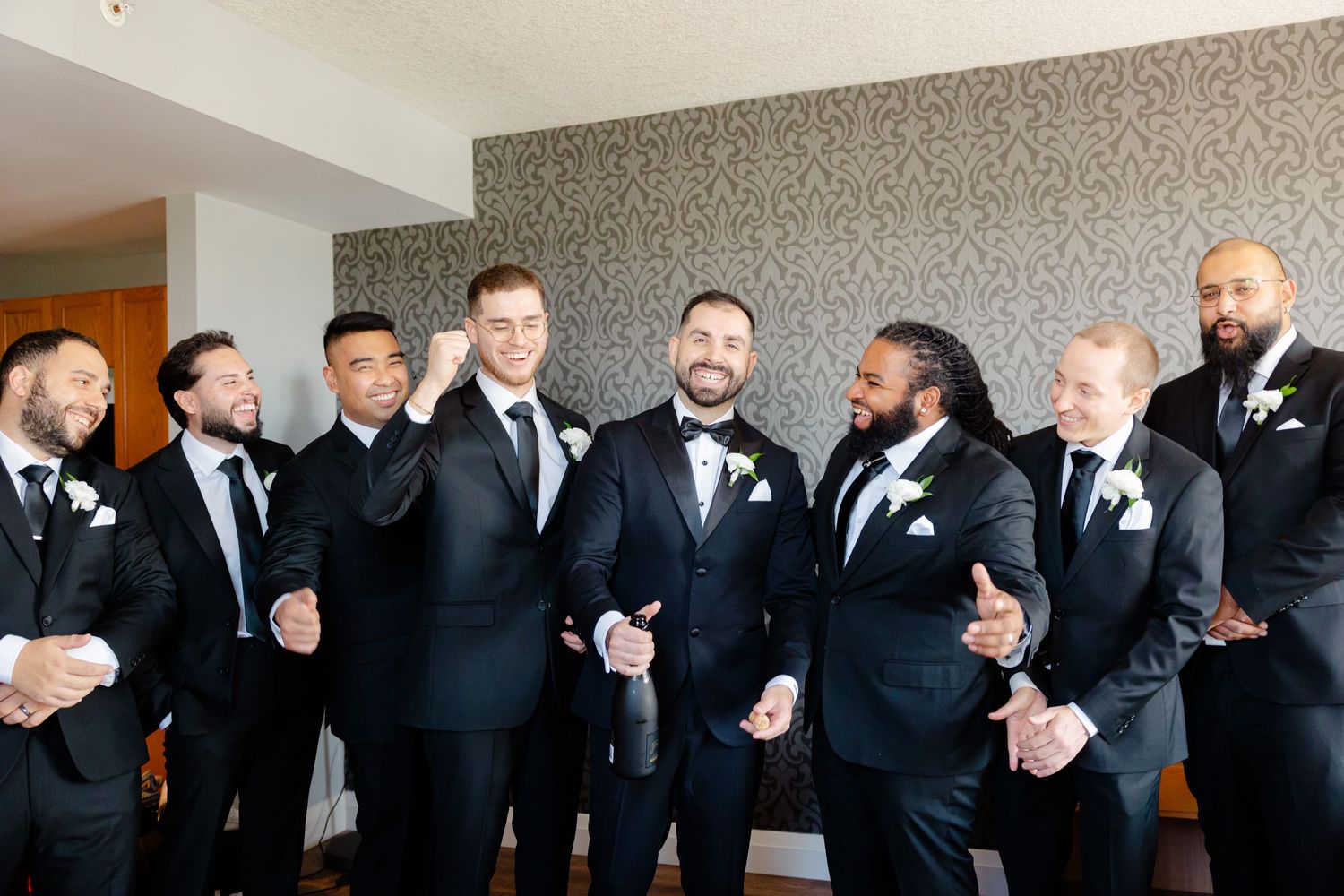 Wedding party in black tuxedos posing together against a modern grey patterned wall.