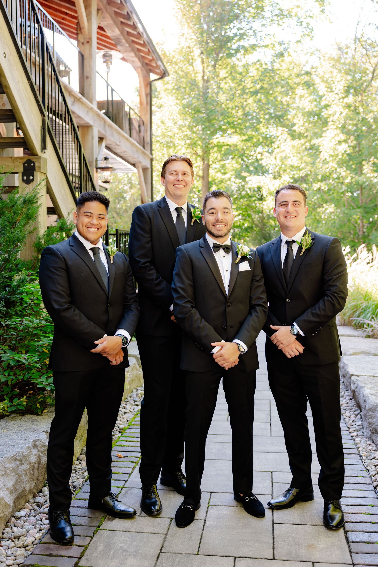 Four groomsmen in formal attire pose together on stone pathway outside rustic building.