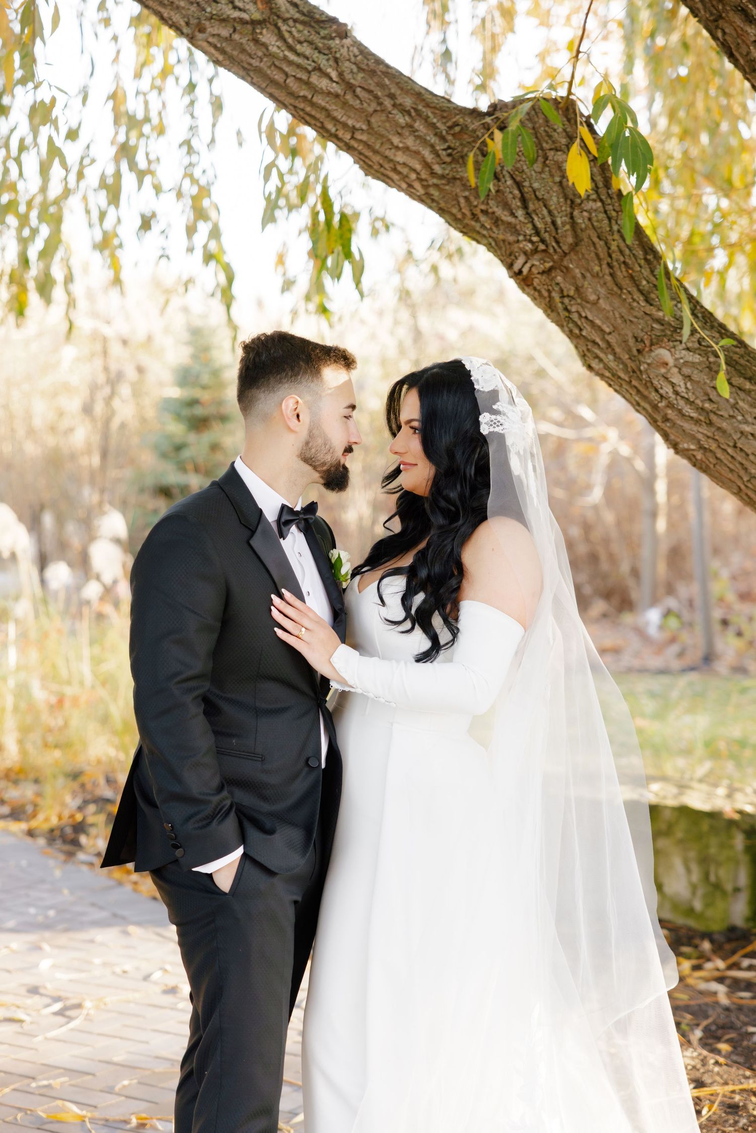 Wedding couple shares tender moment under autumn willow tree on stone path.