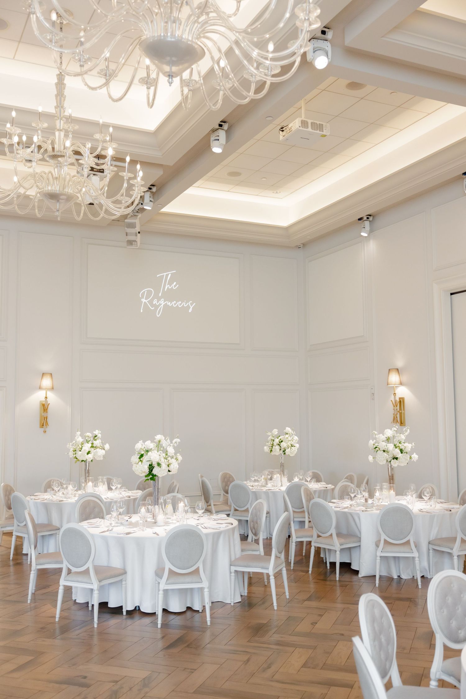 Luxurious white wedding reception room with crystal chandeliers and round tables with elegant place settings.