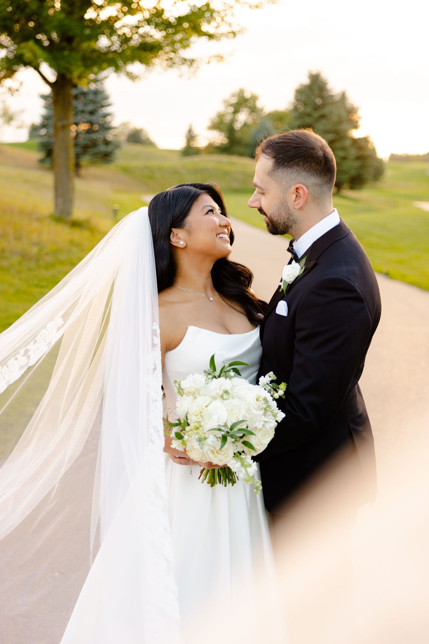 A joyful wedding couple sharing a laugh on a path with flowing veil and white bouquet.